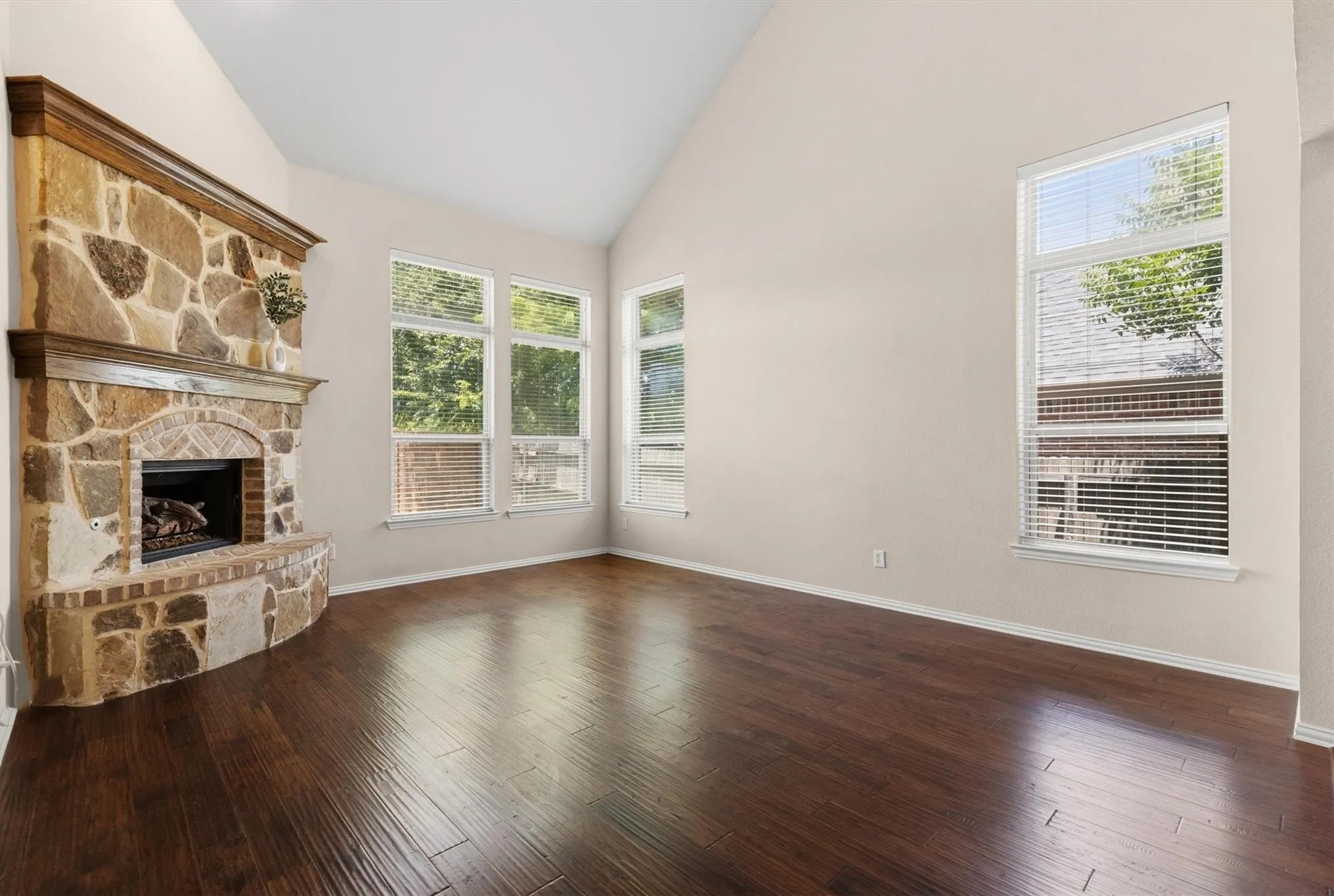 Living space featuring a stone and brick fireplace, vaulted ceiling, rich wood-finish flooring, and multiple windows with blinds