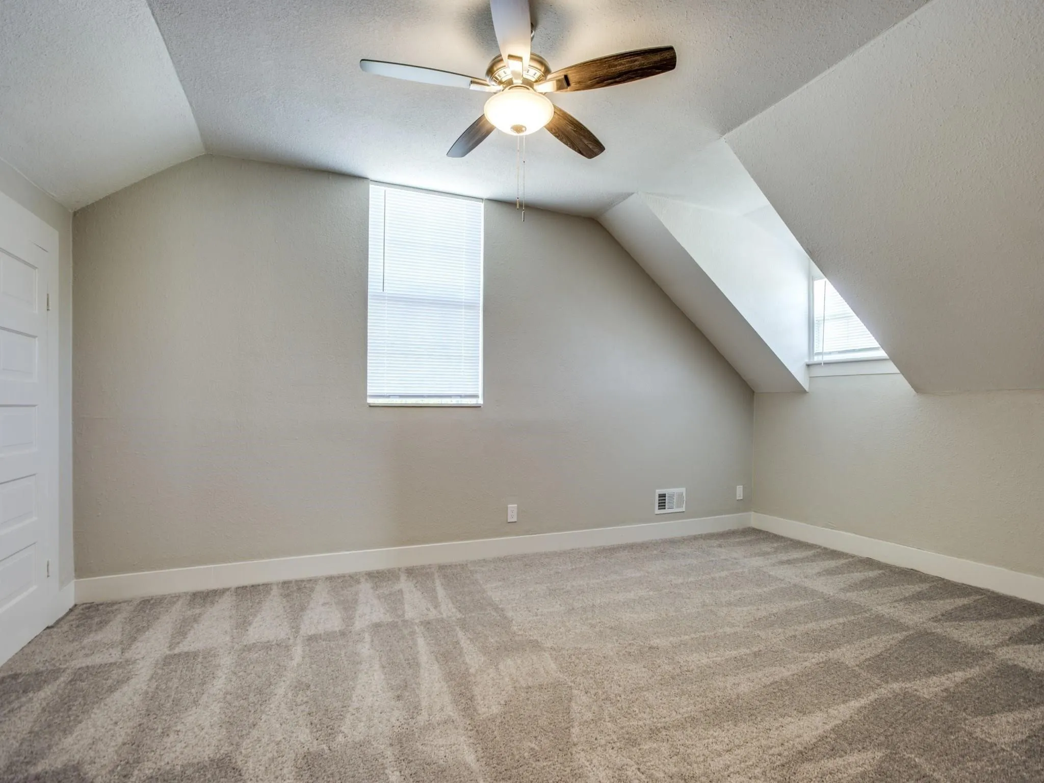 Carpeted room featuring a ceiling fan with light fixture, two windows with blinds, and an angled ceiling design