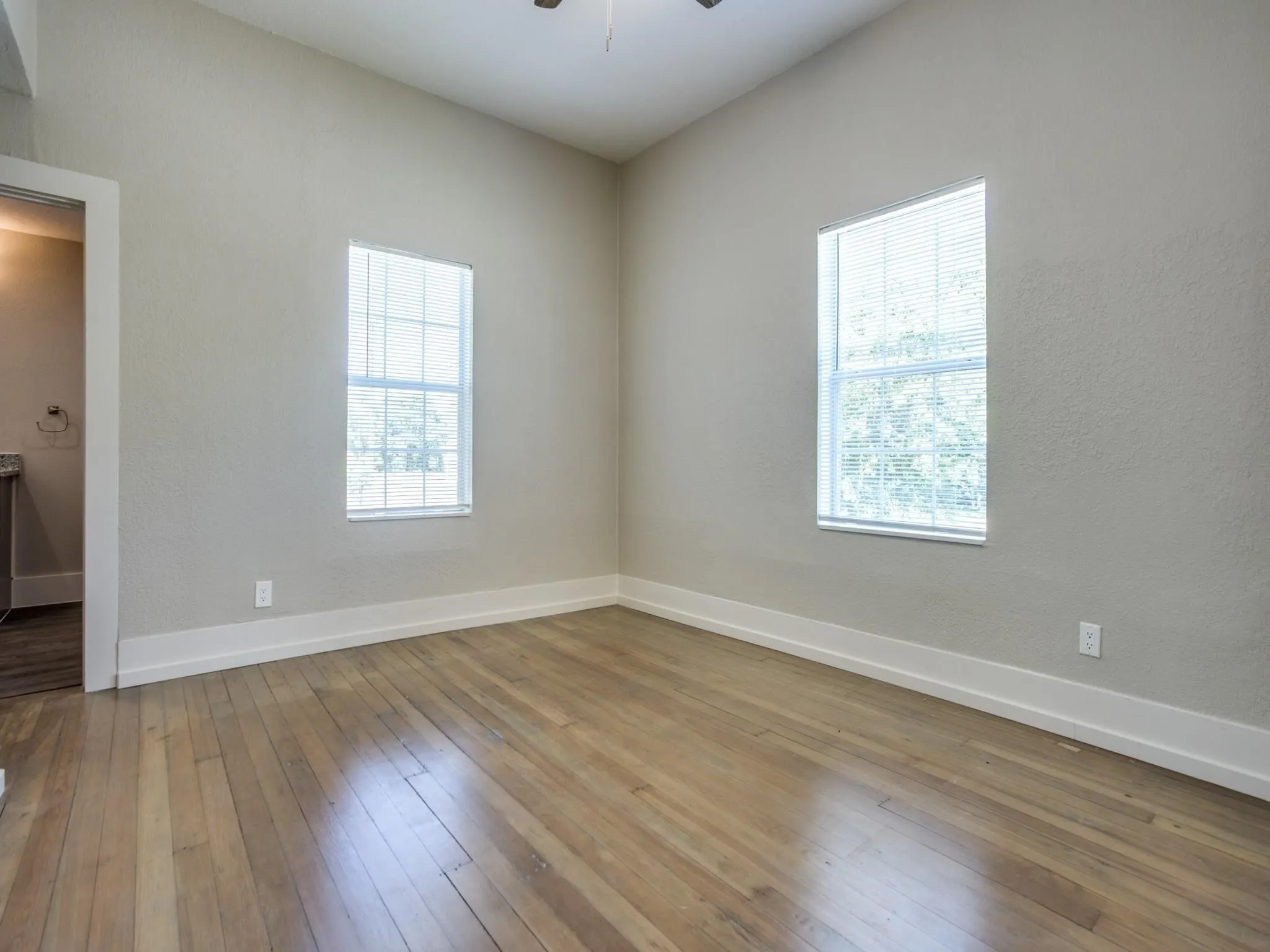 Spacious room featuring wood-finish flooring, light gray walls, two windows with blinds, and white baseboards