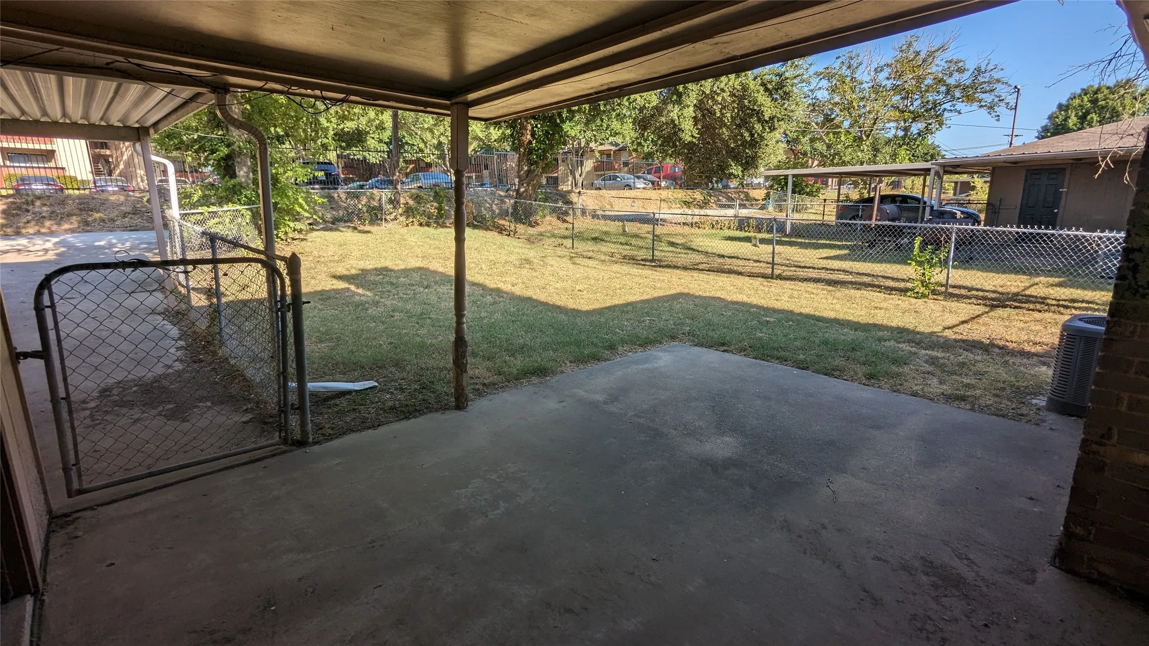 Covered concrete patio opening to an enclosed yard with chain-link fencing, mature trees, and a grassy area