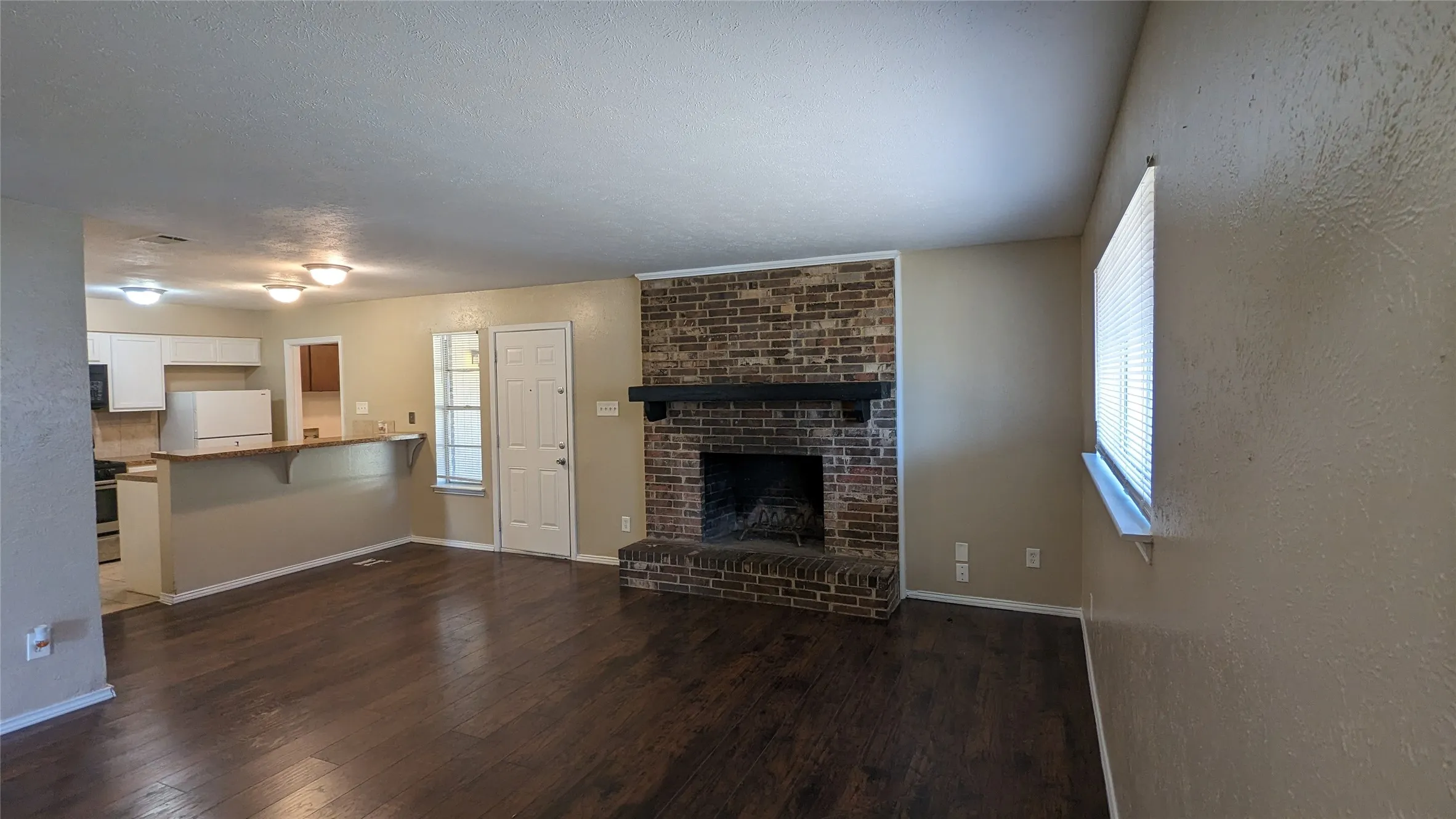 Expansive living area with wood-finish flooring, a prominent brick fireplace with a dark wood mantel, and a kitchen pass-through with a breakfast bar