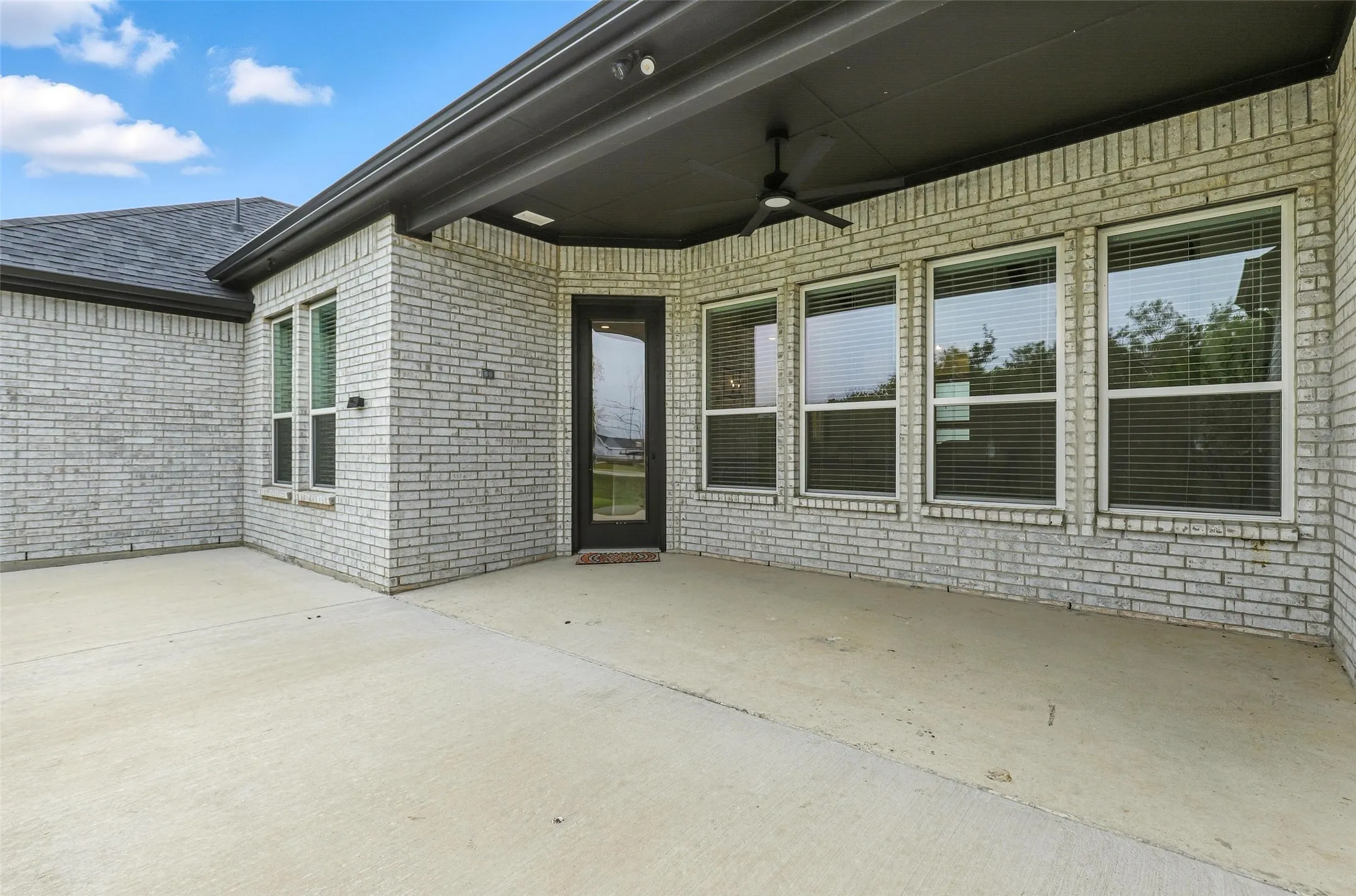 Expansive concrete patio area with a covered ceiling featuring a ceiling fan, white brick exterior, multiple windows, and a glass-paneled door
