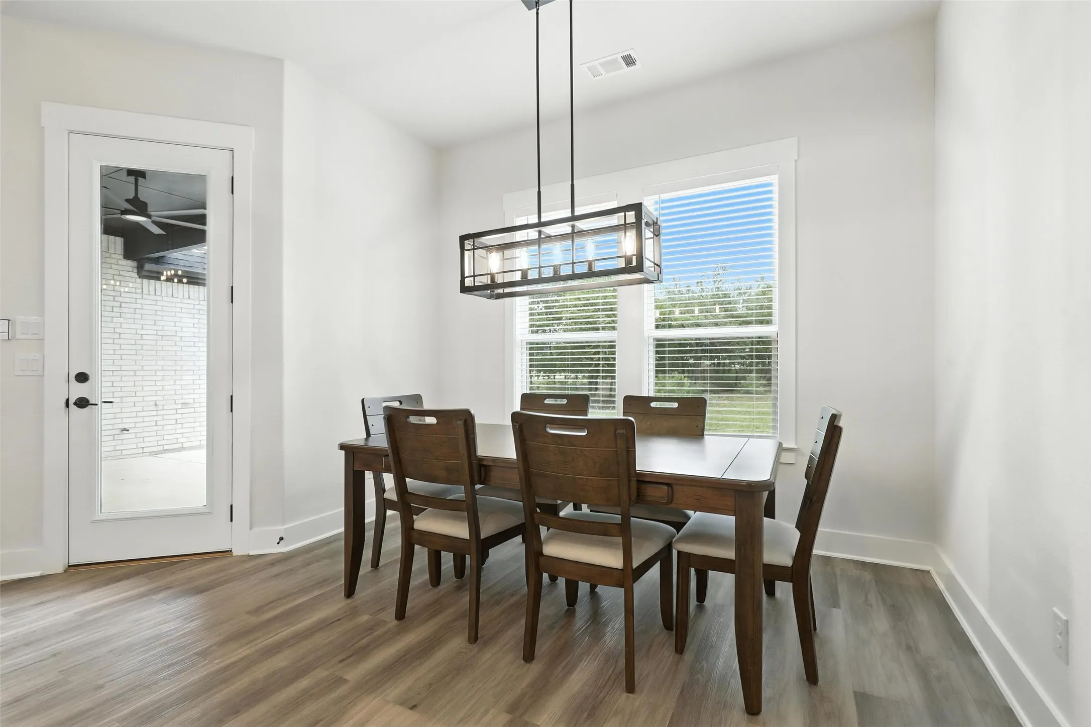 Dining area featuring wood-finish flooring, a contemporary rectangular chandelier, a large window with blinds, and a glass-paneled door leading to an outdoor patio with white brickwork