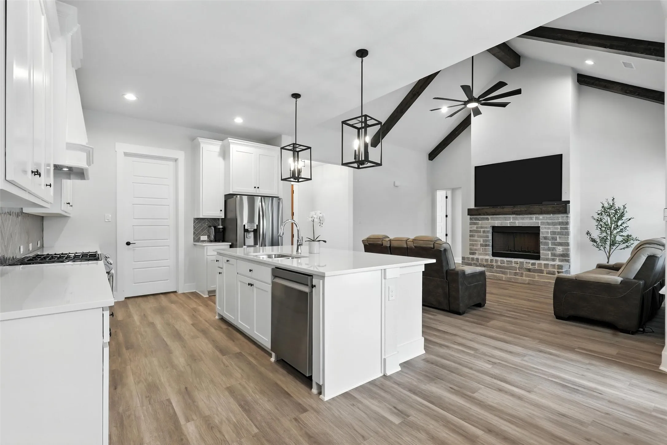 Open-concept living space featuring a kitchen island with sink, stainless steel appliances, wood-finish flooring, a vaulted ceiling with exposed beams, and a stone fireplace