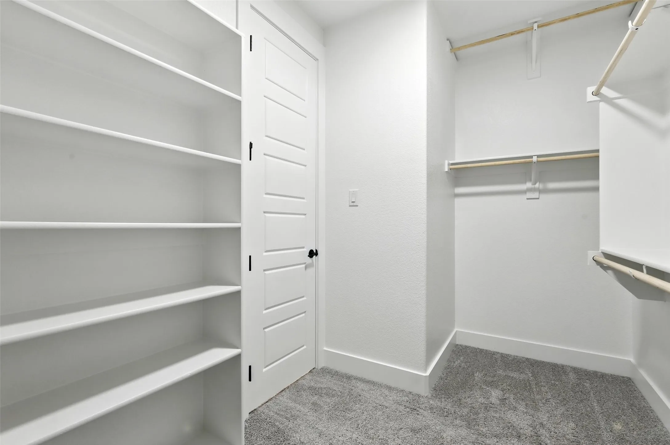 Walk-in closet with extensive built-in shelving, multiple hanging rods, paneled door with matte black hardware, and gray carpet flooring