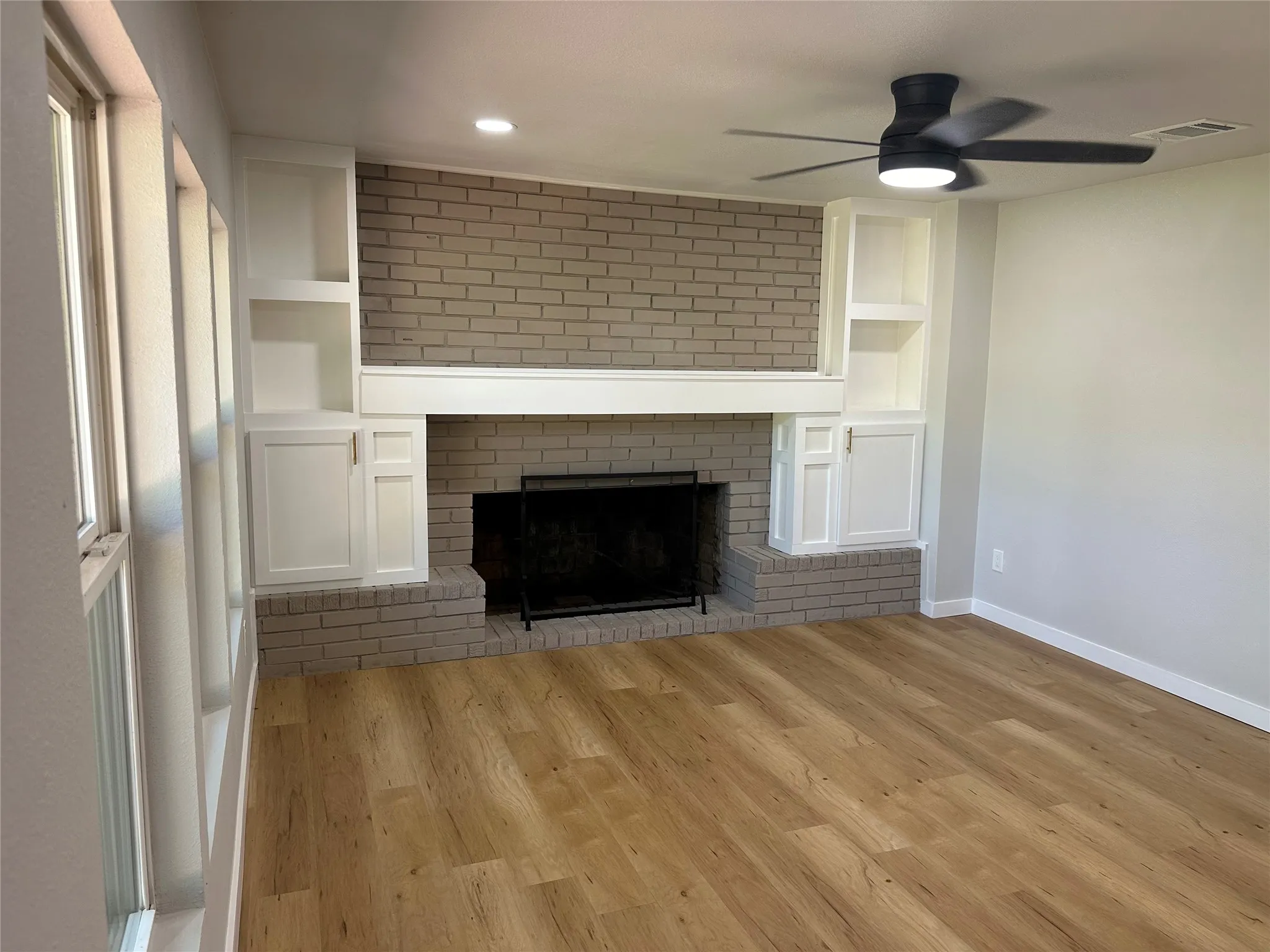 Unfurnished living room with ceiling fan, a fireplace, light wood-type flooring, built in features, and brick wall