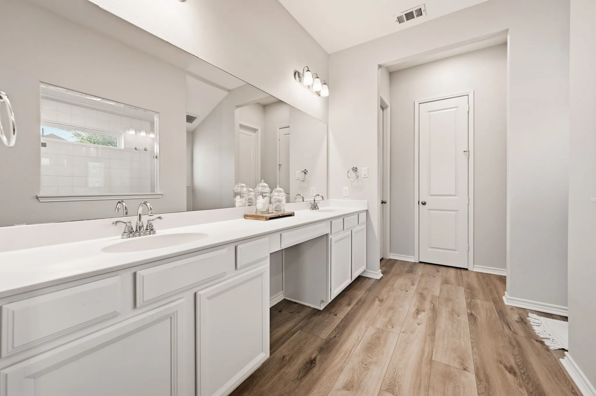 Spacious bathroom featuring an extended double vanity with white cabinetry and integrated sinks, wood-finish flooring, and a large mirror extending the length of the vanity