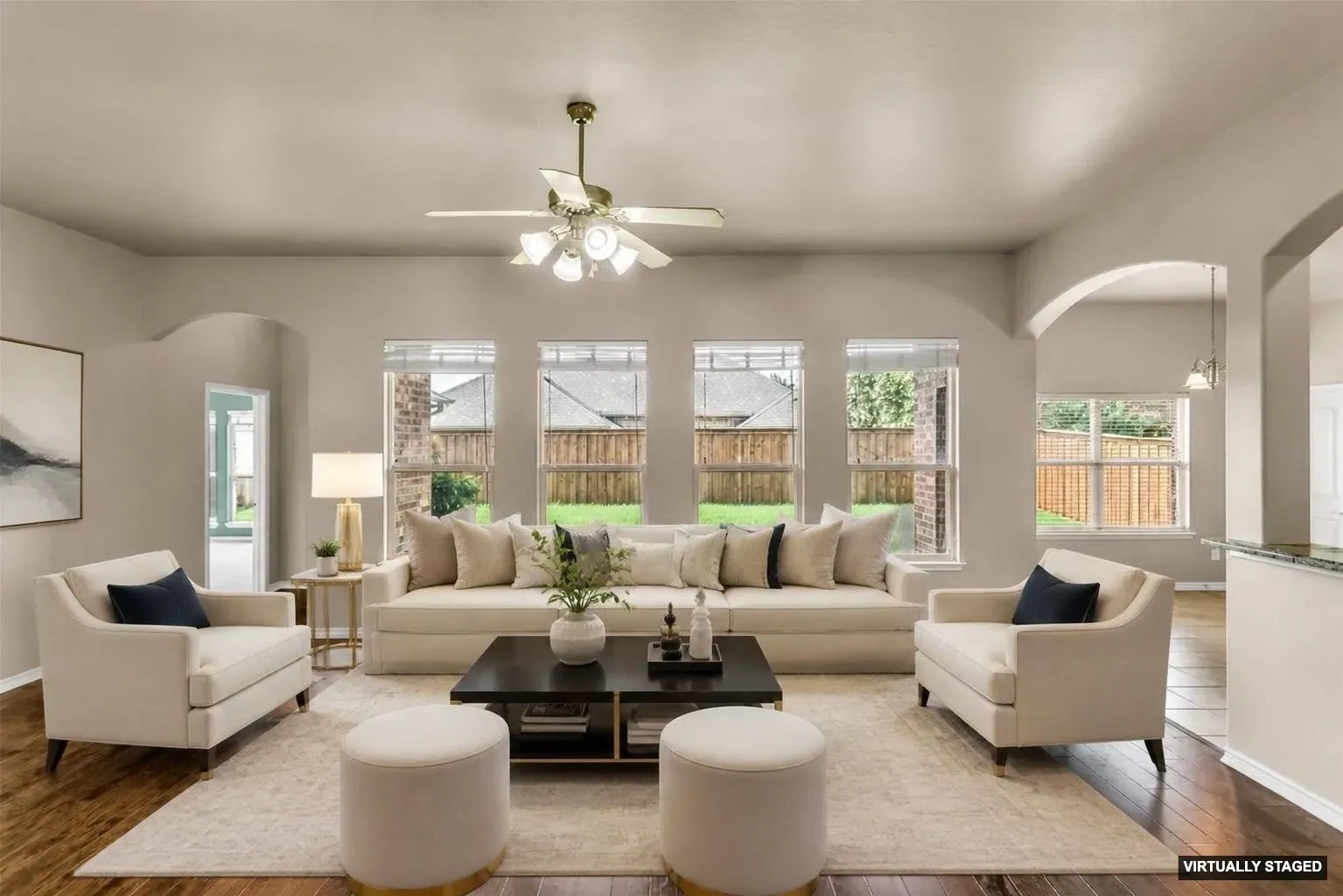 Staged, Living area featuring wood-finish flooring, light-colored walls, arched doorways, a ceiling fan with integrated lighting, and multiple windows overlooking a fenced backyard