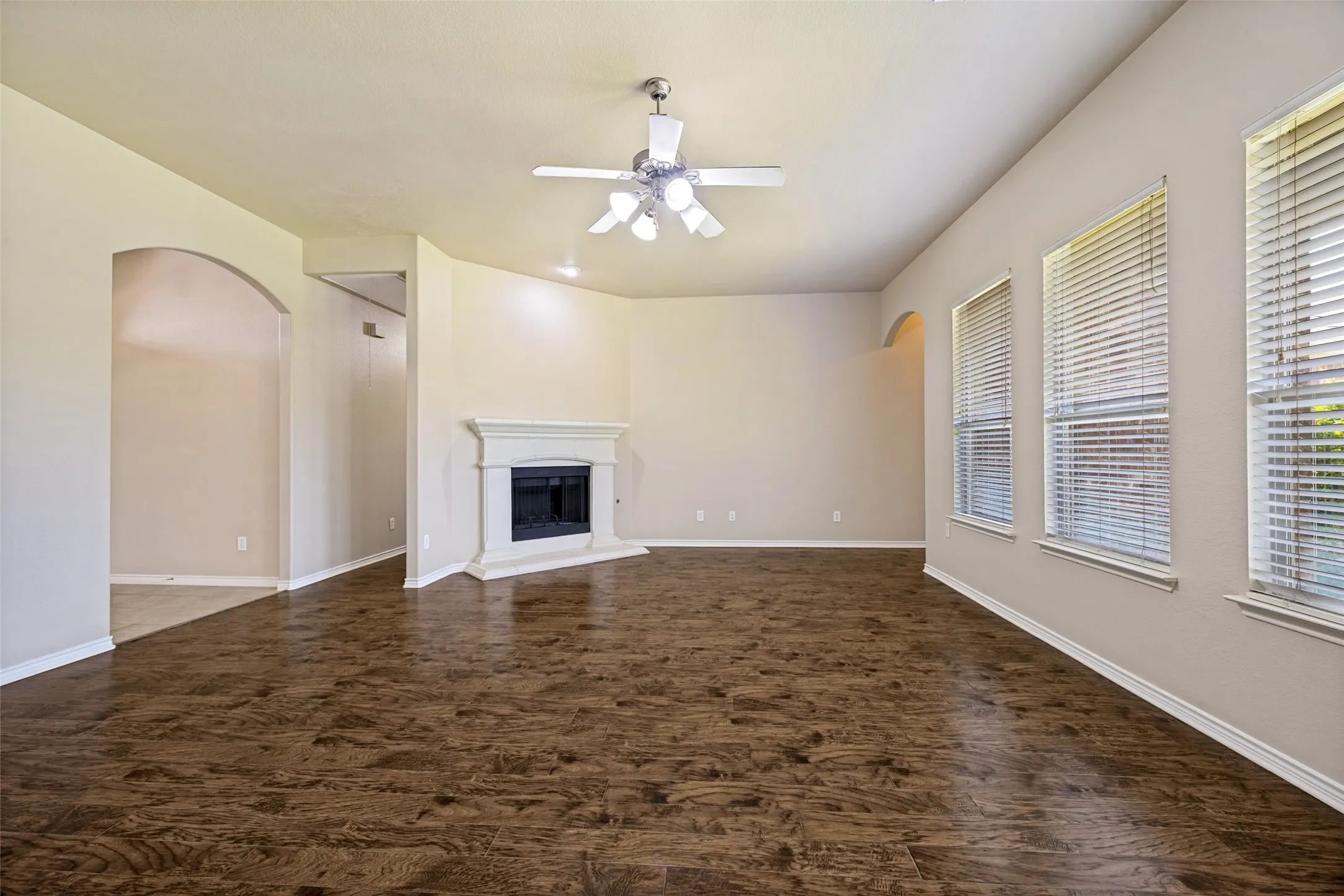 Spacious living area featuring wood-finish flooring, a corner fireplace with a white mantel, multiple windows with blinds, and an overhead ceiling fan