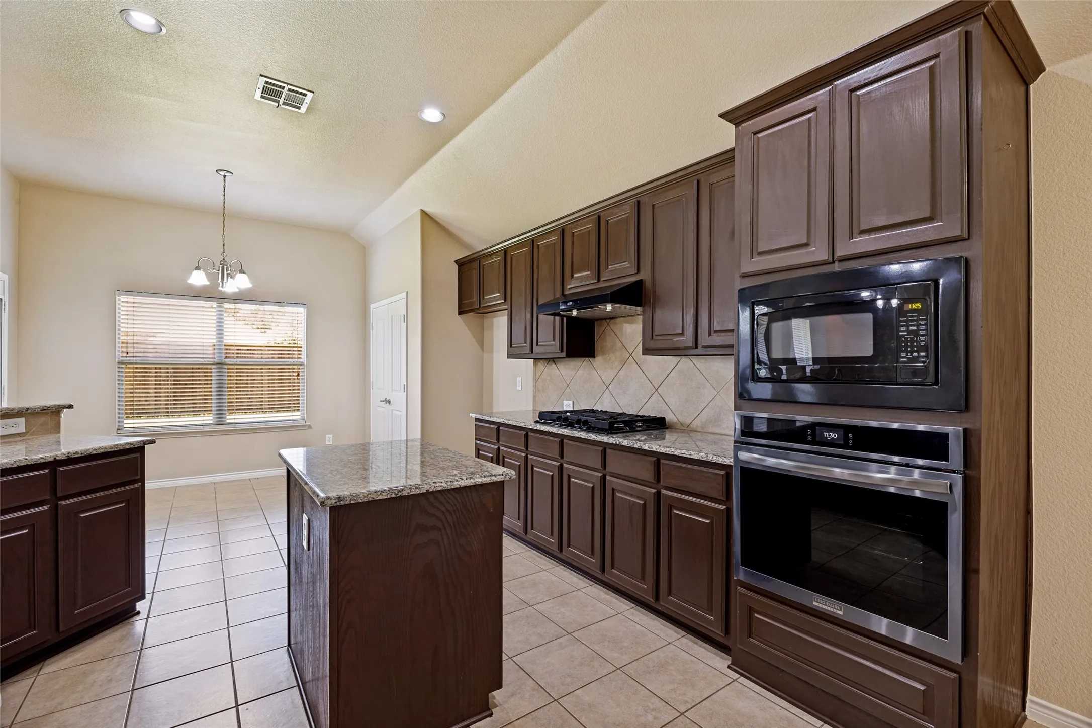 Spacious kitchen featuring dark wood cabinetry, granite countertops, a built-in oven and microwave, a gas cooktop, and a tile backsplash