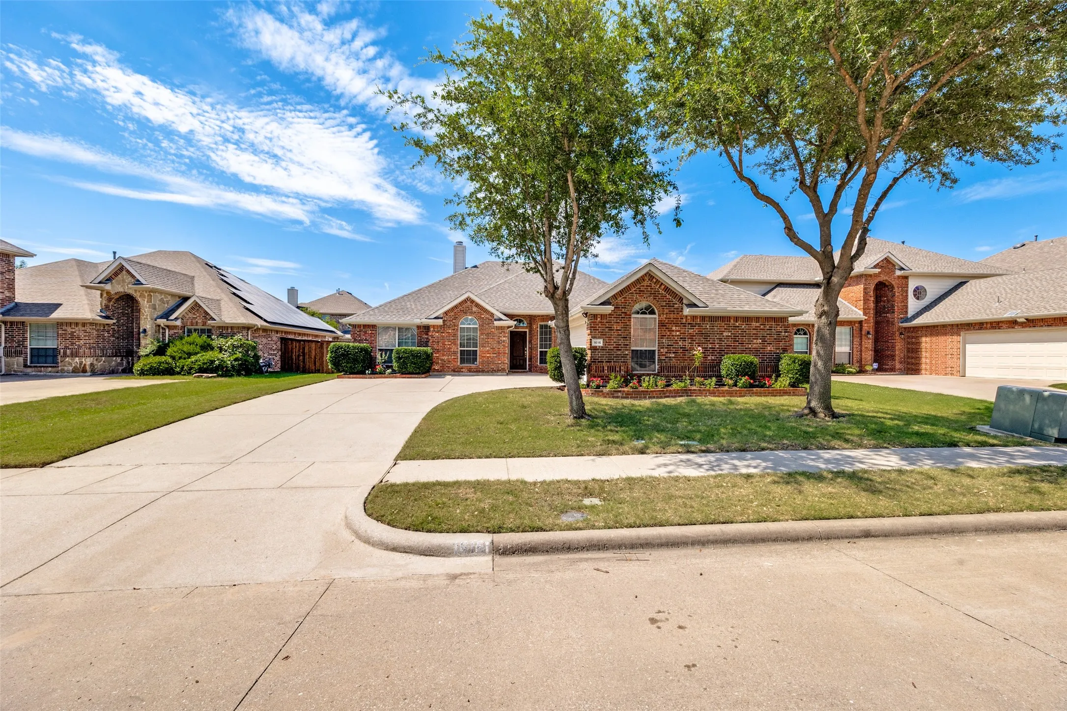 Brick facade residence featuring an arched entry, a concrete driveway, an attached garage, landscaped garden beds, and mature trees