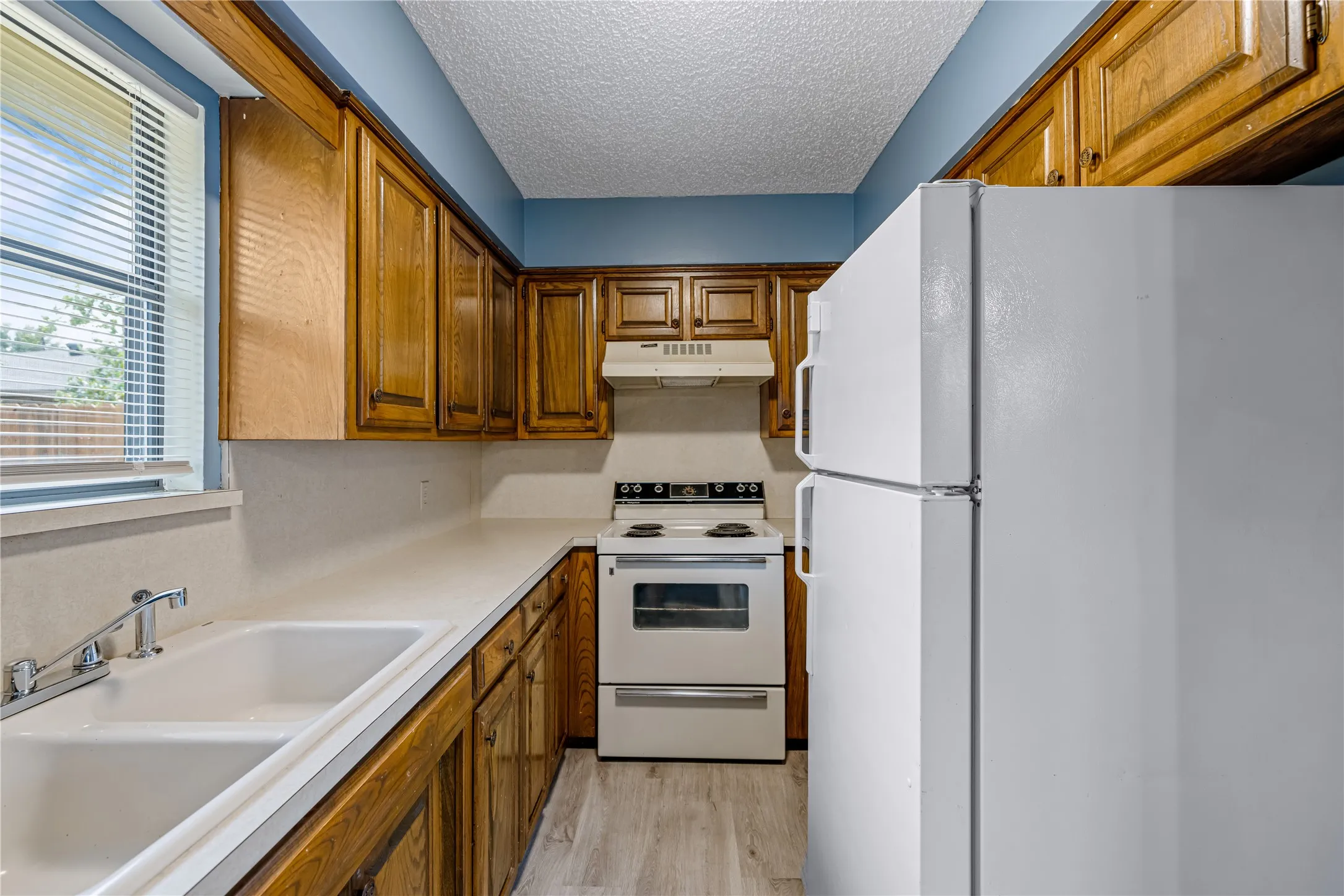Kitchen featuring under cabinet range hood, brown cabinetry, white appliances, a textured ceiling, and a sink