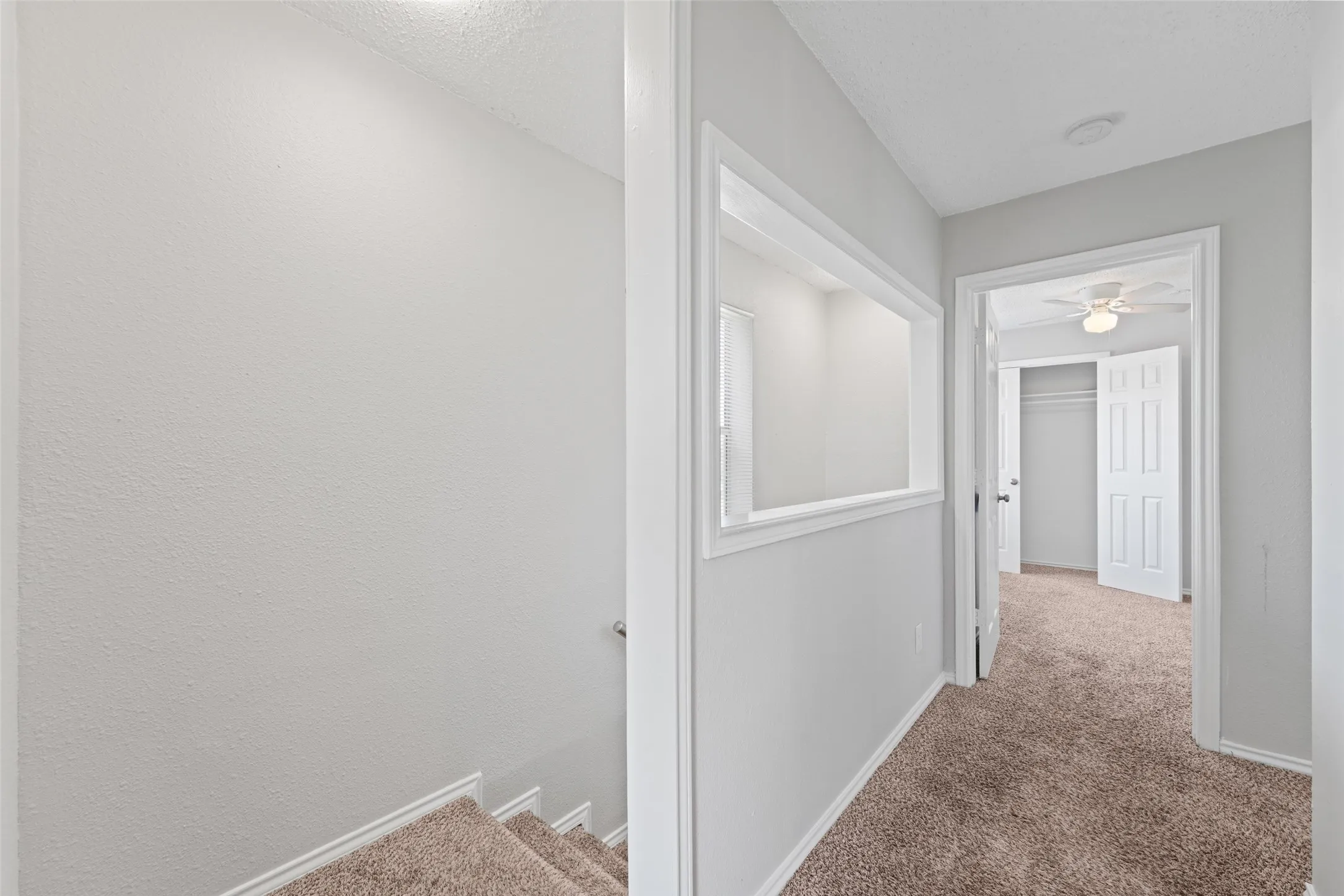 Carpeted hallway with a wall cutout feature and paneled doors, illuminated by recessed lighting and a ceiling fan in the distance