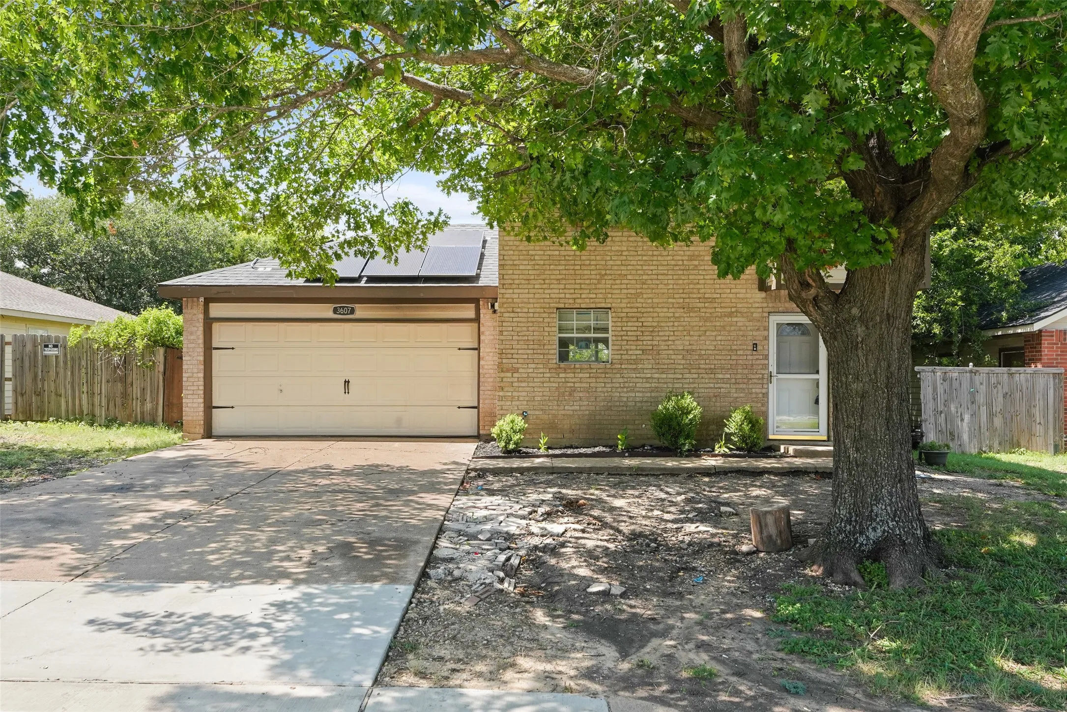 Brick exterior featuring a single-car garage with a panel door, solar panels on the roof, a prominent mature tree, a paved driveway, and a fenced side yard