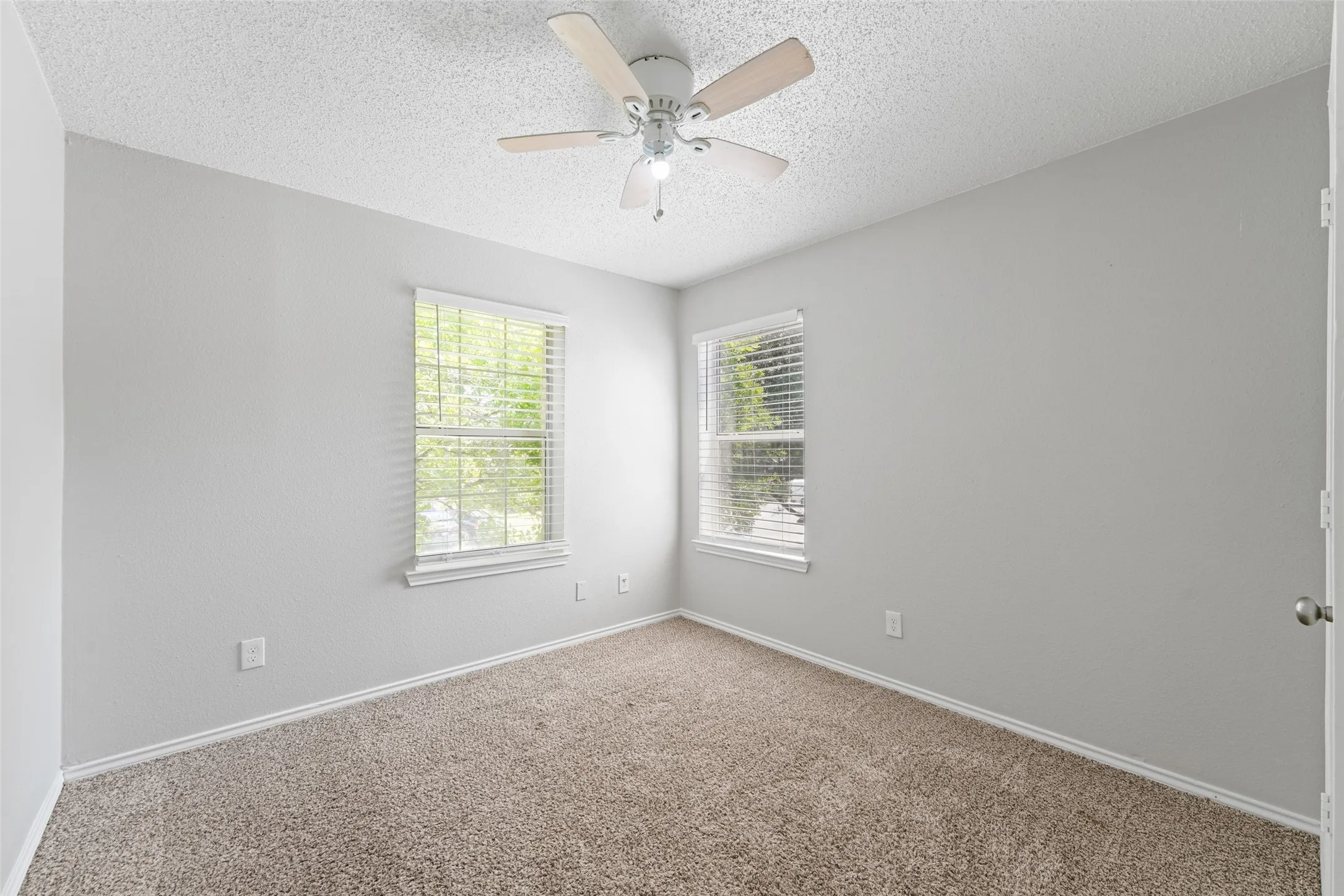 Carpeted room featuring a ceiling fan with light fixture, two windows with horizontal blinds, textured walls, and white baseboards