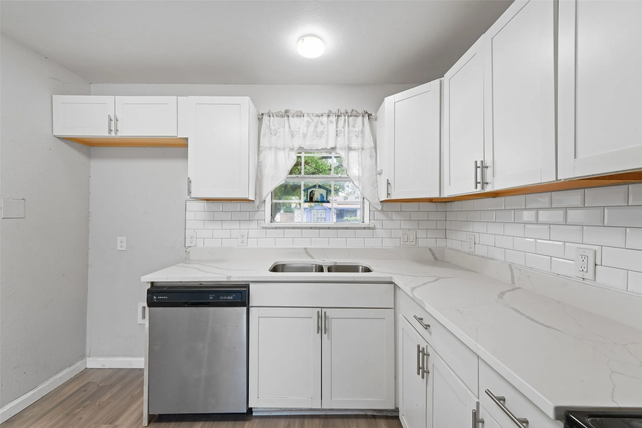 Kitchen featuring white shaker cabinetry, light-toned countertops with subtle veining, a stainless steel dishwasher, a double basin sink, and wood-finish flooring