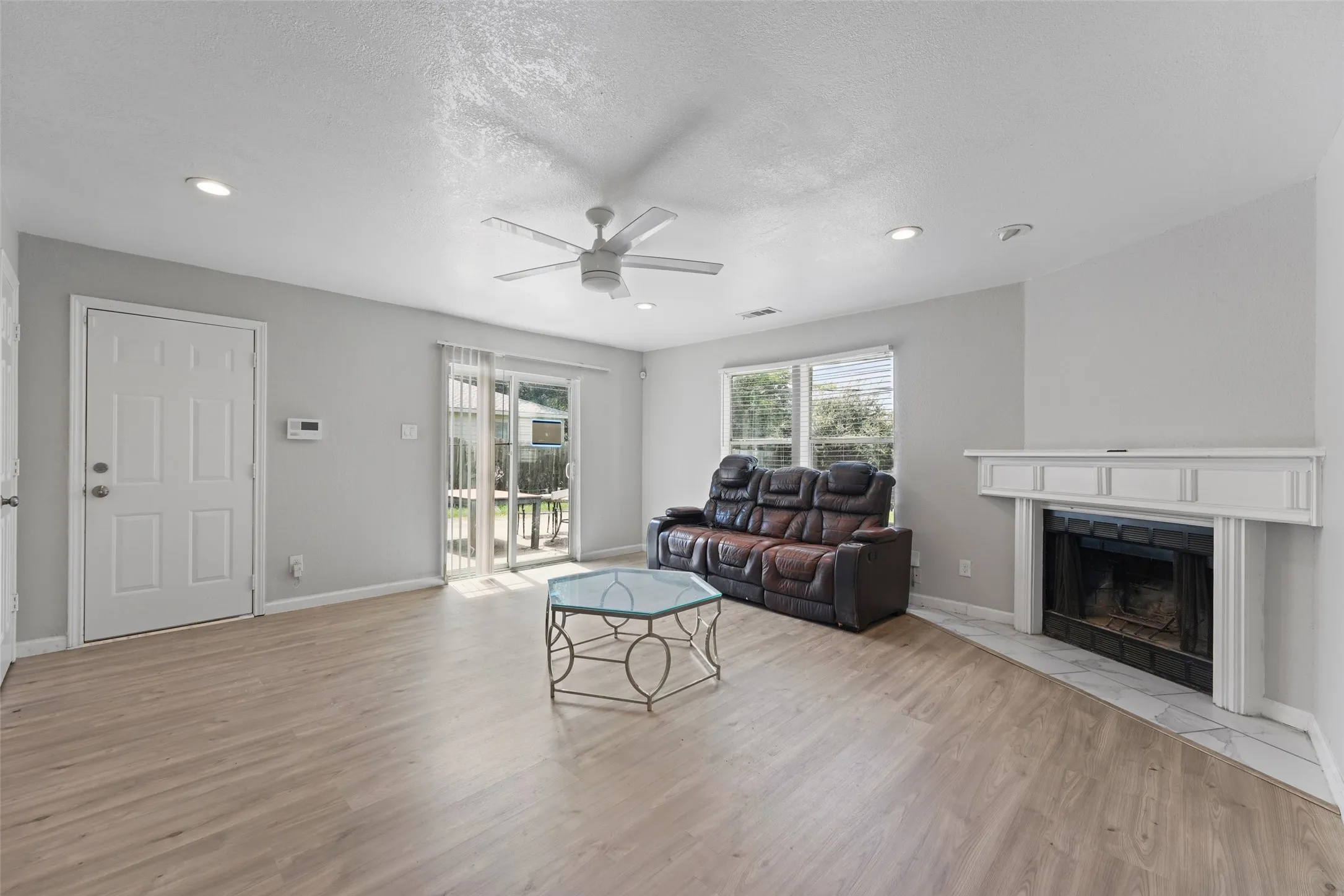 Spacious living area featuring light wood-finish flooring, a white trim fireplace, ceiling fan, recessed lighting, and sliding glass door access to an outdoor area