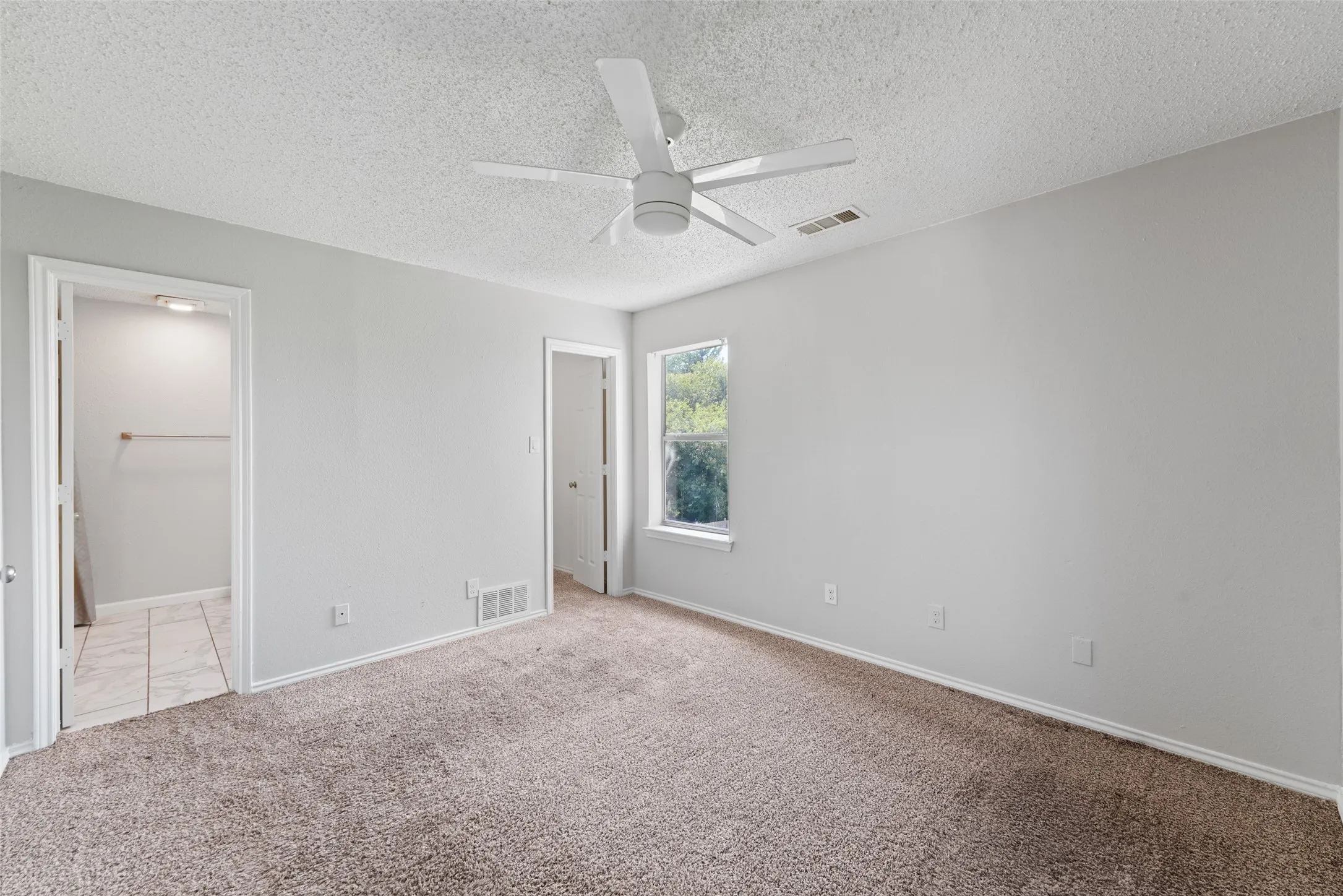 Spacious room featuring neutral carpeting, light gray walls, a ceiling fan with integrated lighting, a window providing natural light, and an adjacent walk-in closet with tile flooring