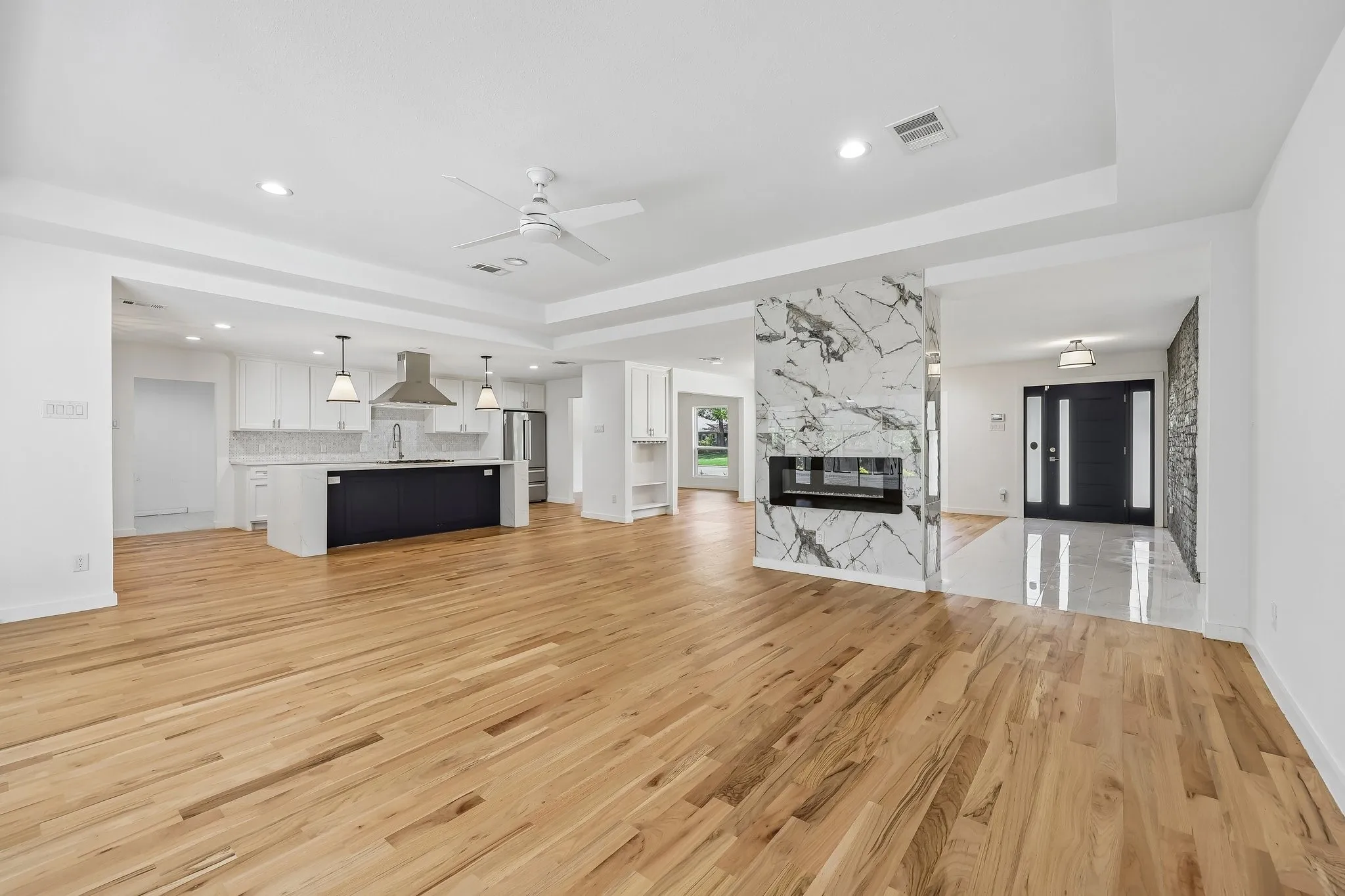 Open-concept living space with wood-finish flooring, a contemporary fireplace with a marble-style surround, a kitchen featuring white cabinetry and a dark island with pendant lighting, and recessed ceiling lights