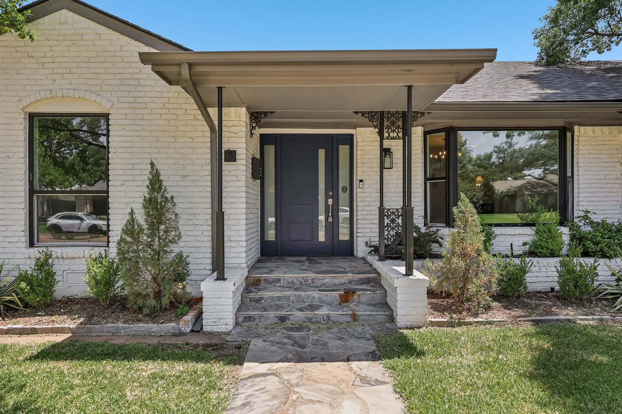 White painted brick exterior featuring a dark blue entry door with sidelights, a covered porch, stone slab pathway, and a bay window