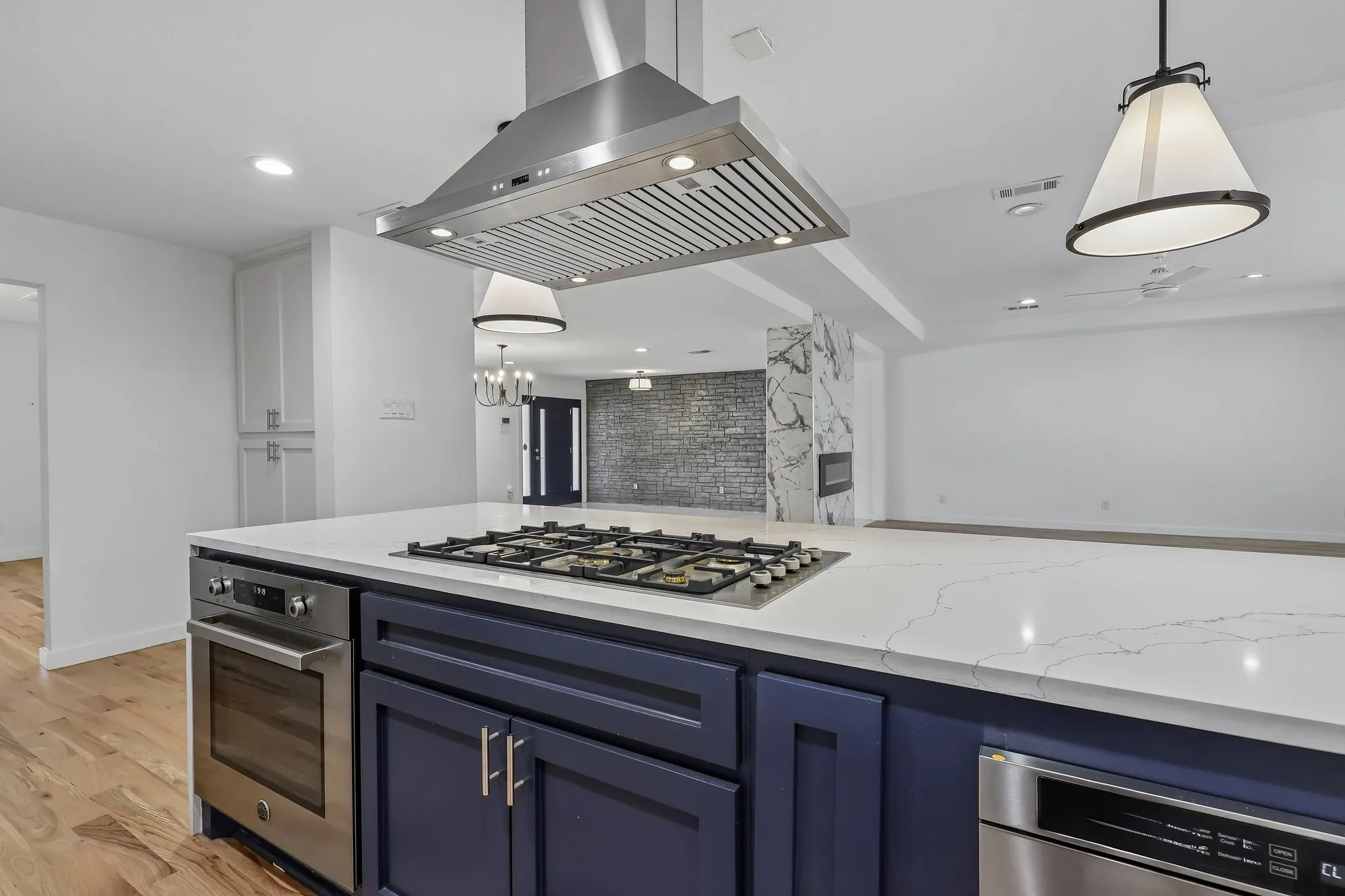 Kitchen island featuring a built-in gas cooktop with an overhead stainless steel range hood, integrated oven, and dishwasher