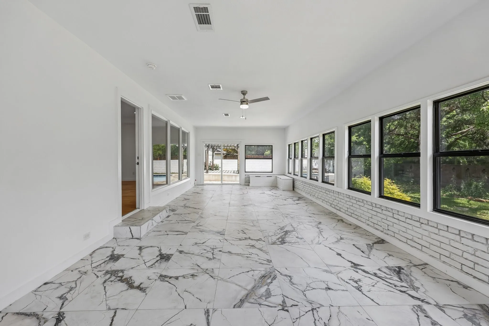 Spacious sunroom featuring large format porcelain tile flooring, a white painted brick accent wall, multiple large windows, and a ceiling fan