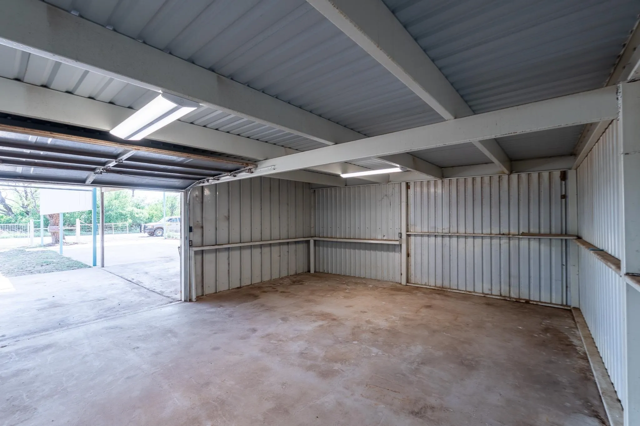 Garage with concrete flooring, corrugated metal walls, exposed structural beams, and overhead fluorescent lighting