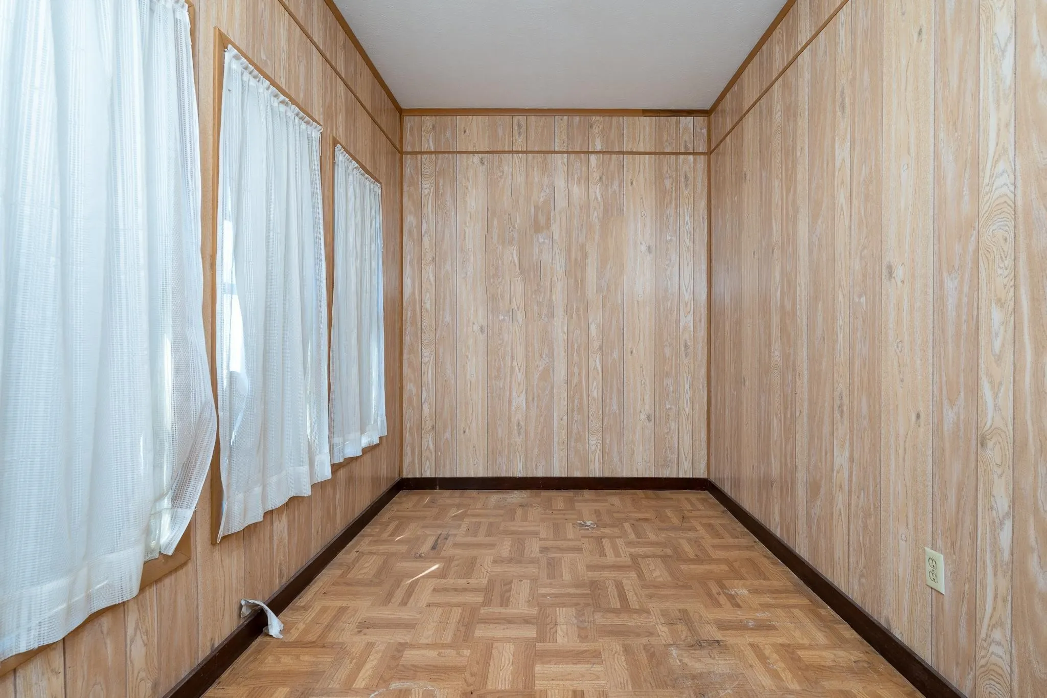 Room featuring wood paneling, parquet flooring, and three windows with sheer curtains