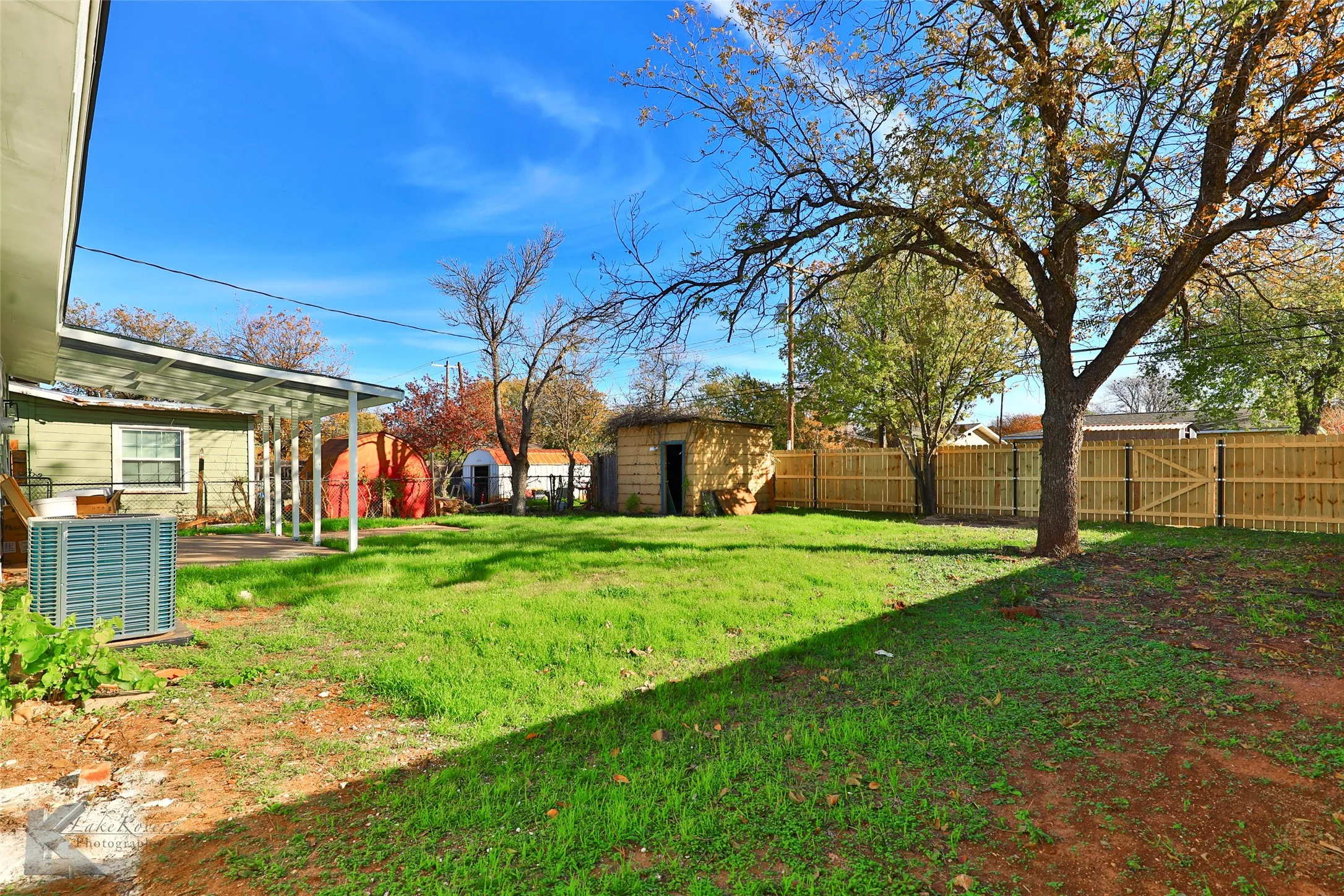 Expansive fenced backyard featuring a large mature tree, grass lawn, and a covered patio area