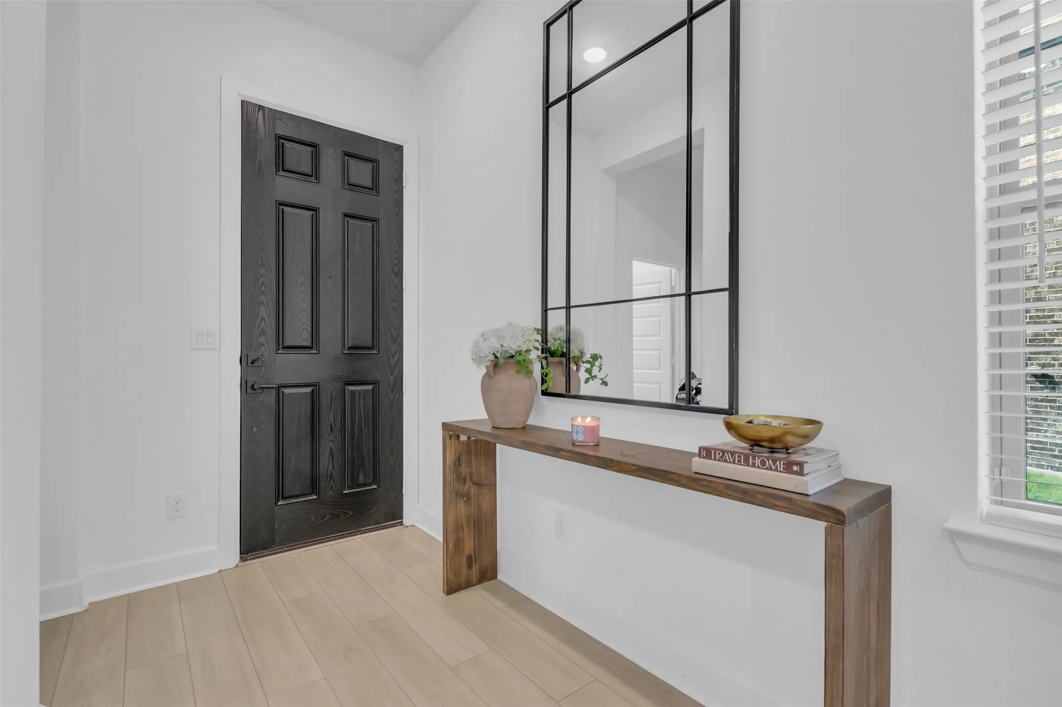 Entryway featuring a dark wood-finish paneled door, light wood-finish flooring, a contemporary console table, and a large black-framed grid mirror