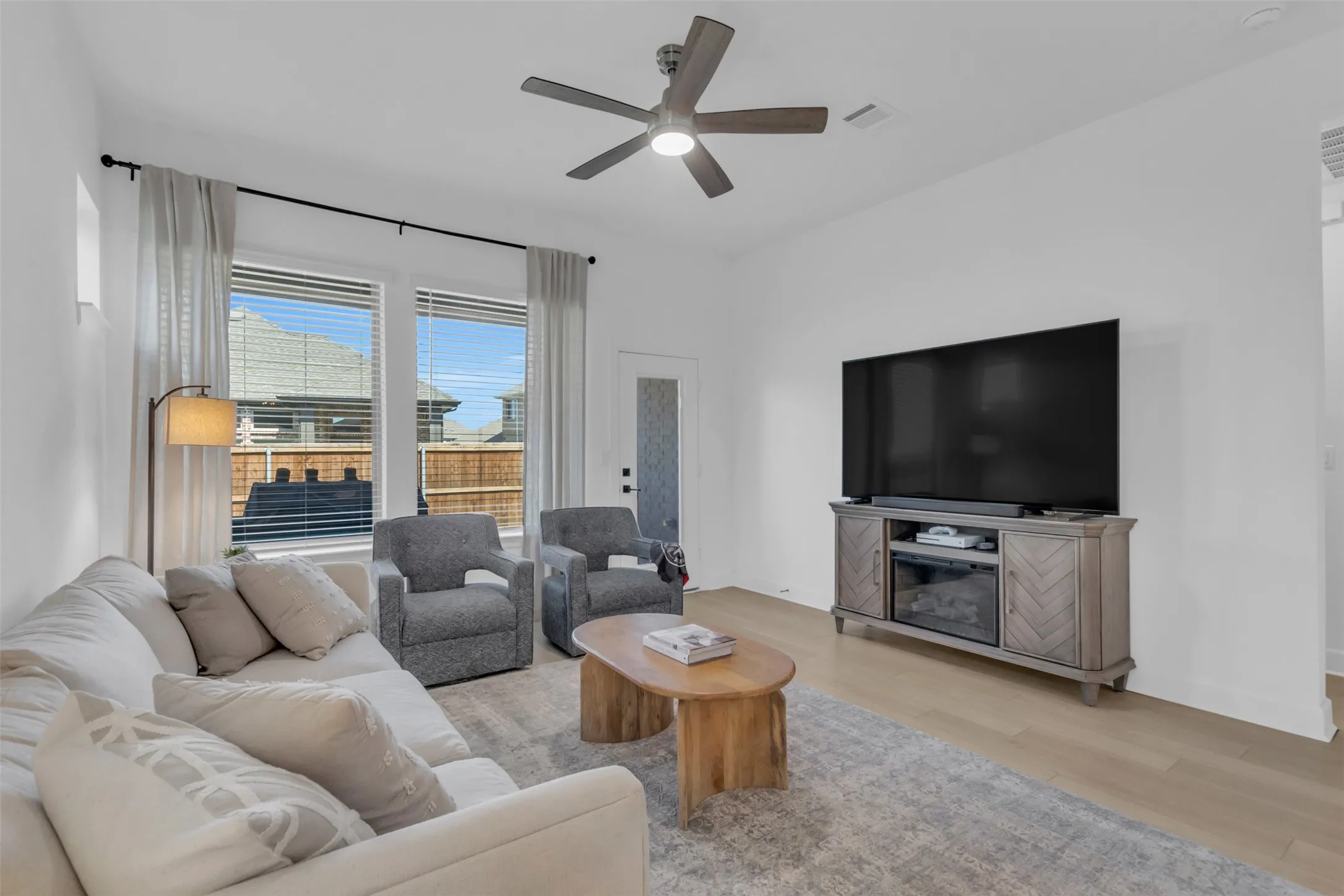 Spacious living area featuring wood-finish flooring, a ceiling fan with integrated lighting, and multiple large windows with horizontal blinds