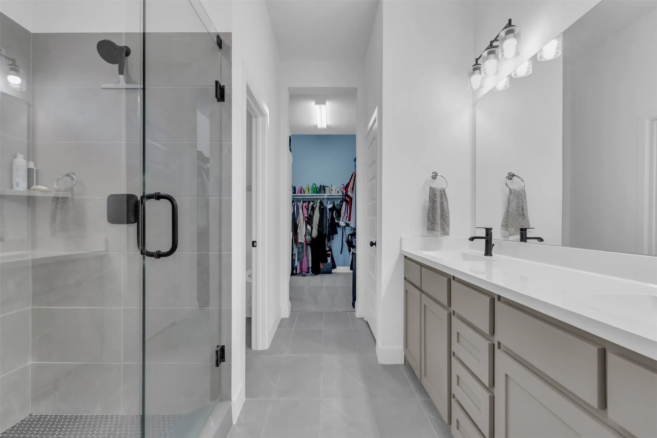 Bathroom featuring a glass-enclosed shower with matte black fixtures, a dual vanity with a white countertop and dark faucets, a large mirror, and tiled flooring