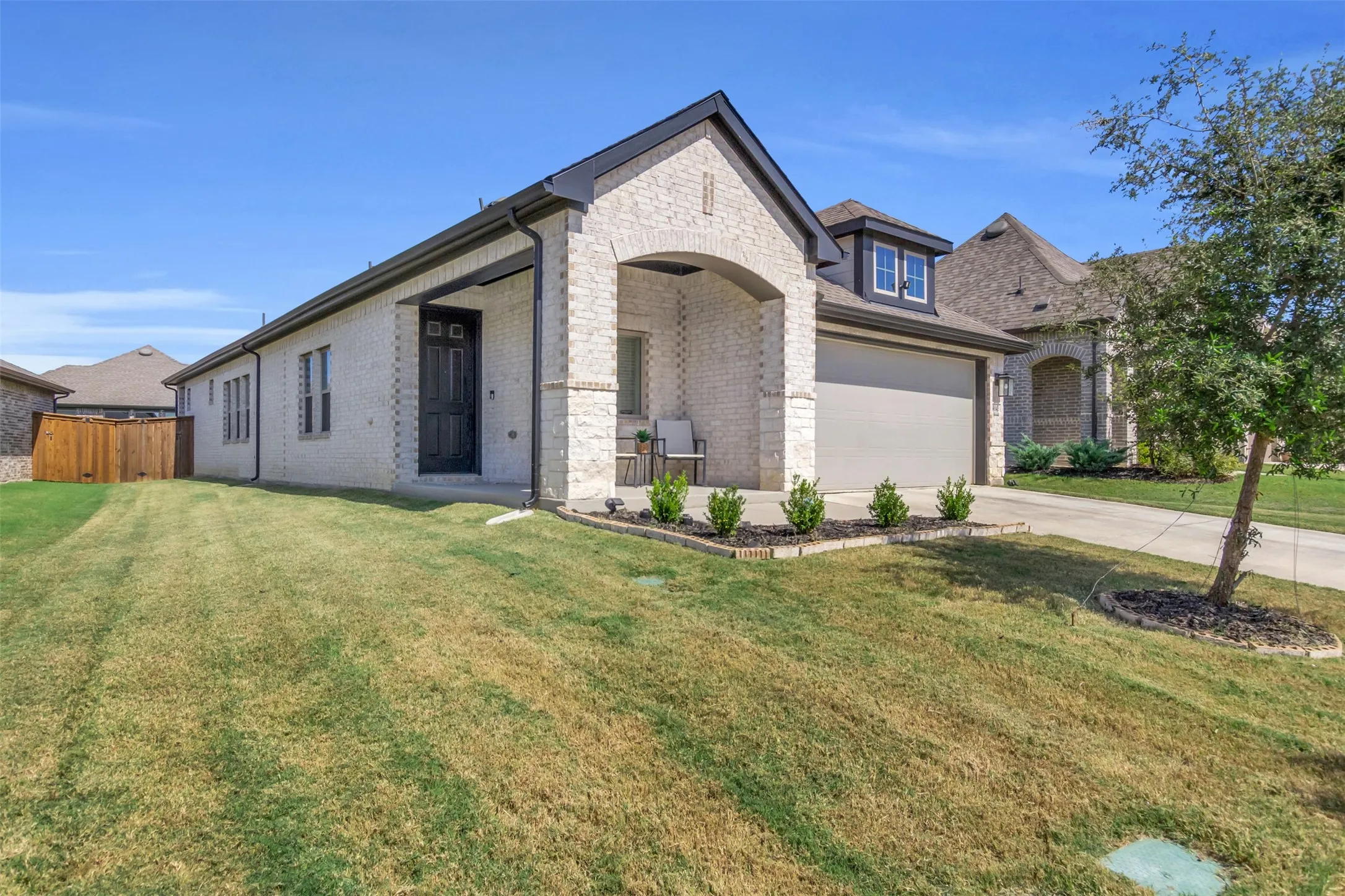 Contemporary single-story residence featuring light-colored brick veneer, a two-car garage, a covered entry porch, dark trim accents, and a dormer window above the garage