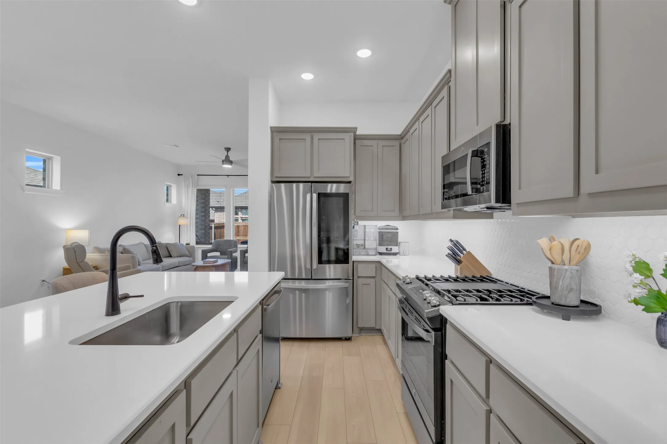 Contemporary kitchen featuring an island with an undermount sink and matte black faucet, light gray cabinetry, stainless steel appliances, white countertops, and a full tile backsplash