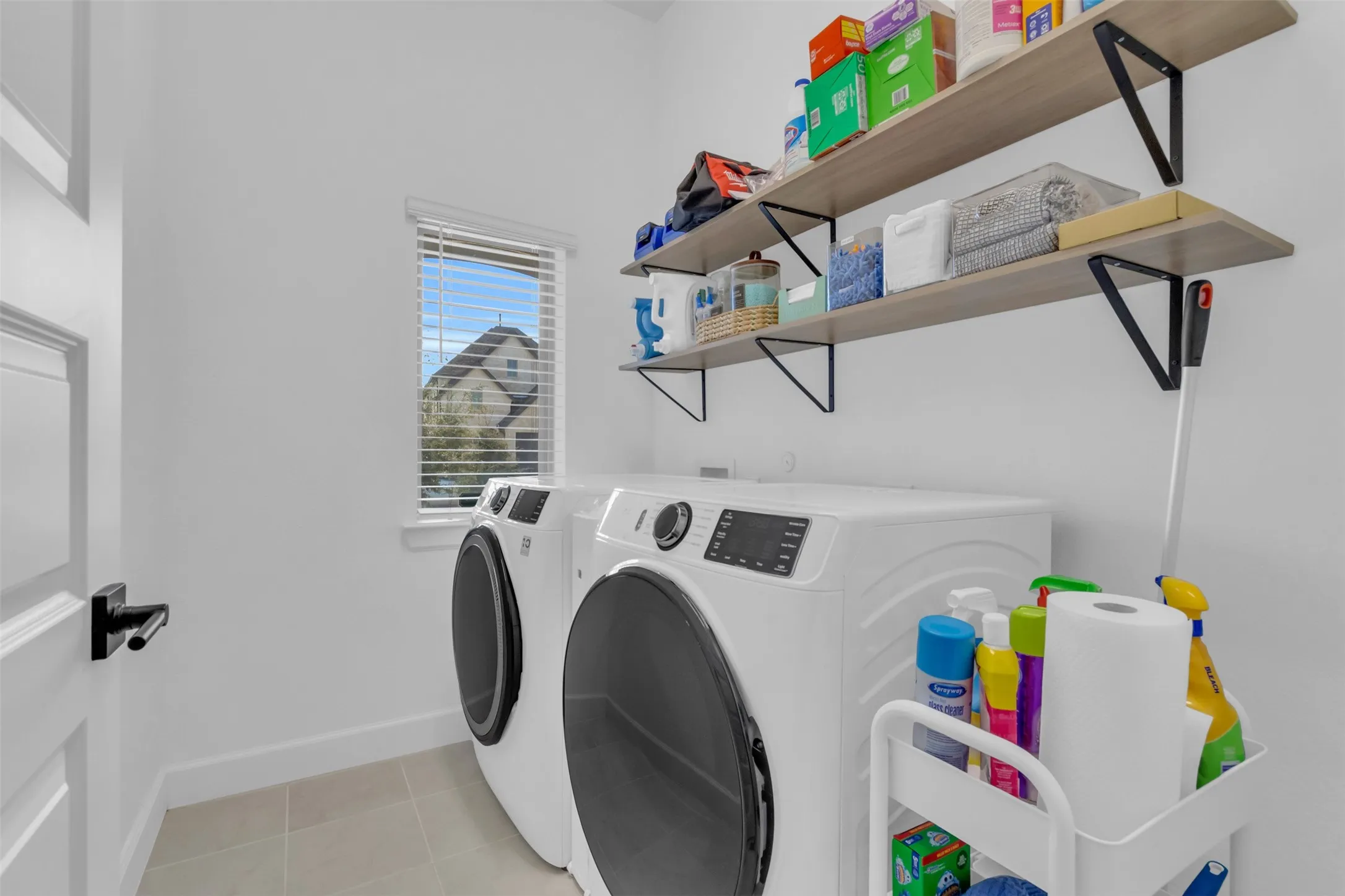 Dedicated laundry space featuring a white front-load washer and dryer set, two wood-finish floating shelves with black brackets, a single window with horizontal blinds, and neutral tile flooring