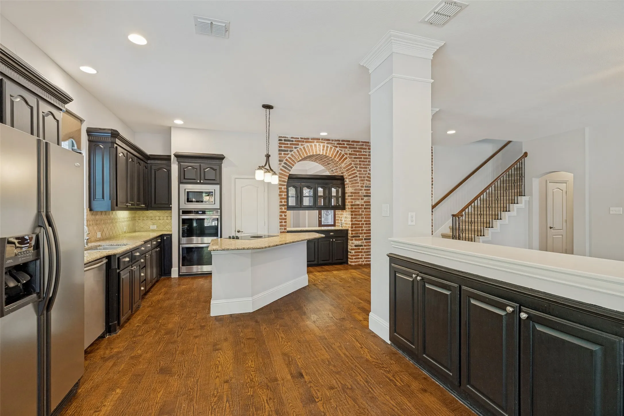 Kitchen featuring dark wood-finish cabinetry, stainless steel appliances, a center island with a light-toned countertop, wood-finish flooring, and a brick archway