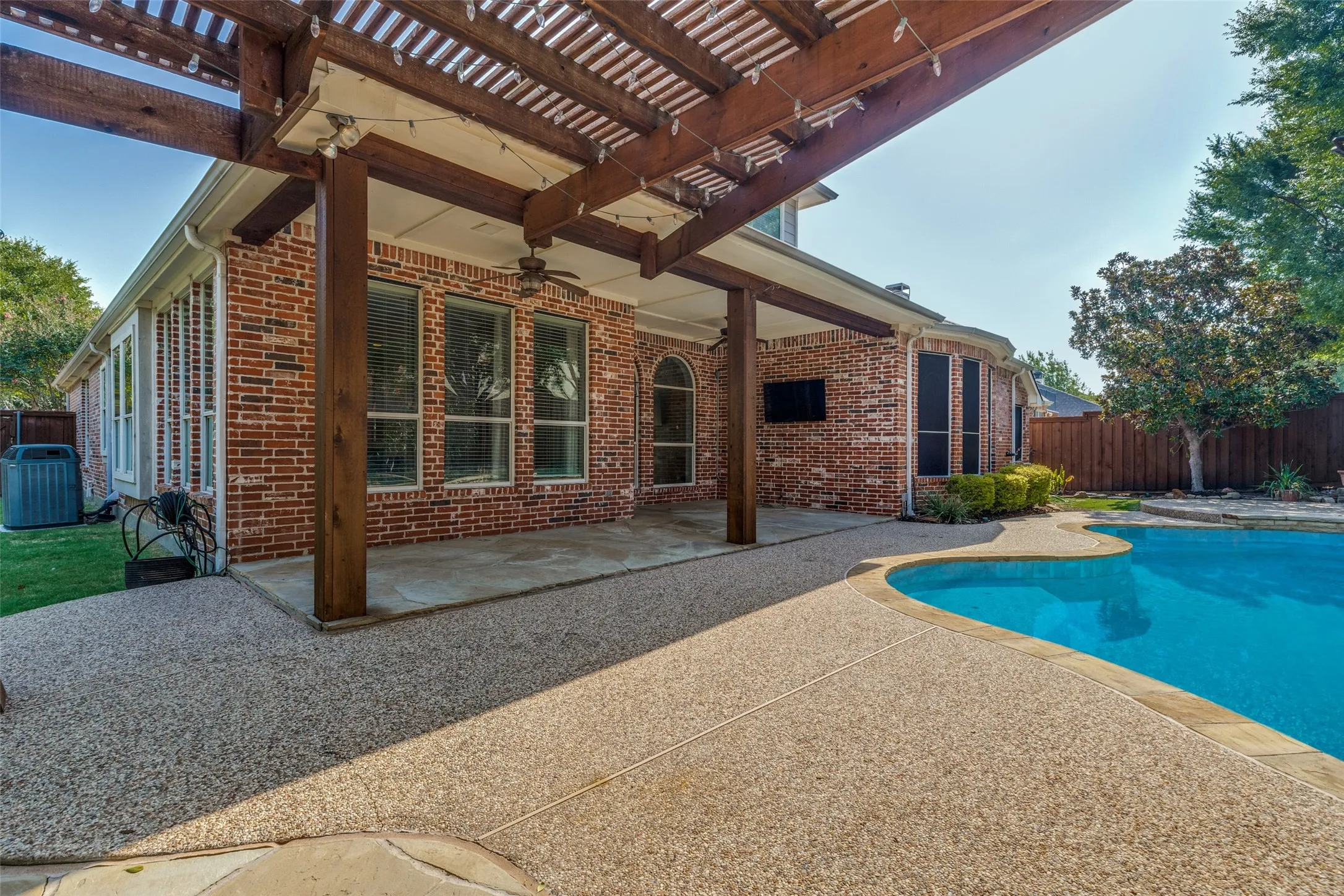 Brick exterior with a covered patio, wood pergola, built-in ceiling fan, and an integrated swimming pool with a textured deck