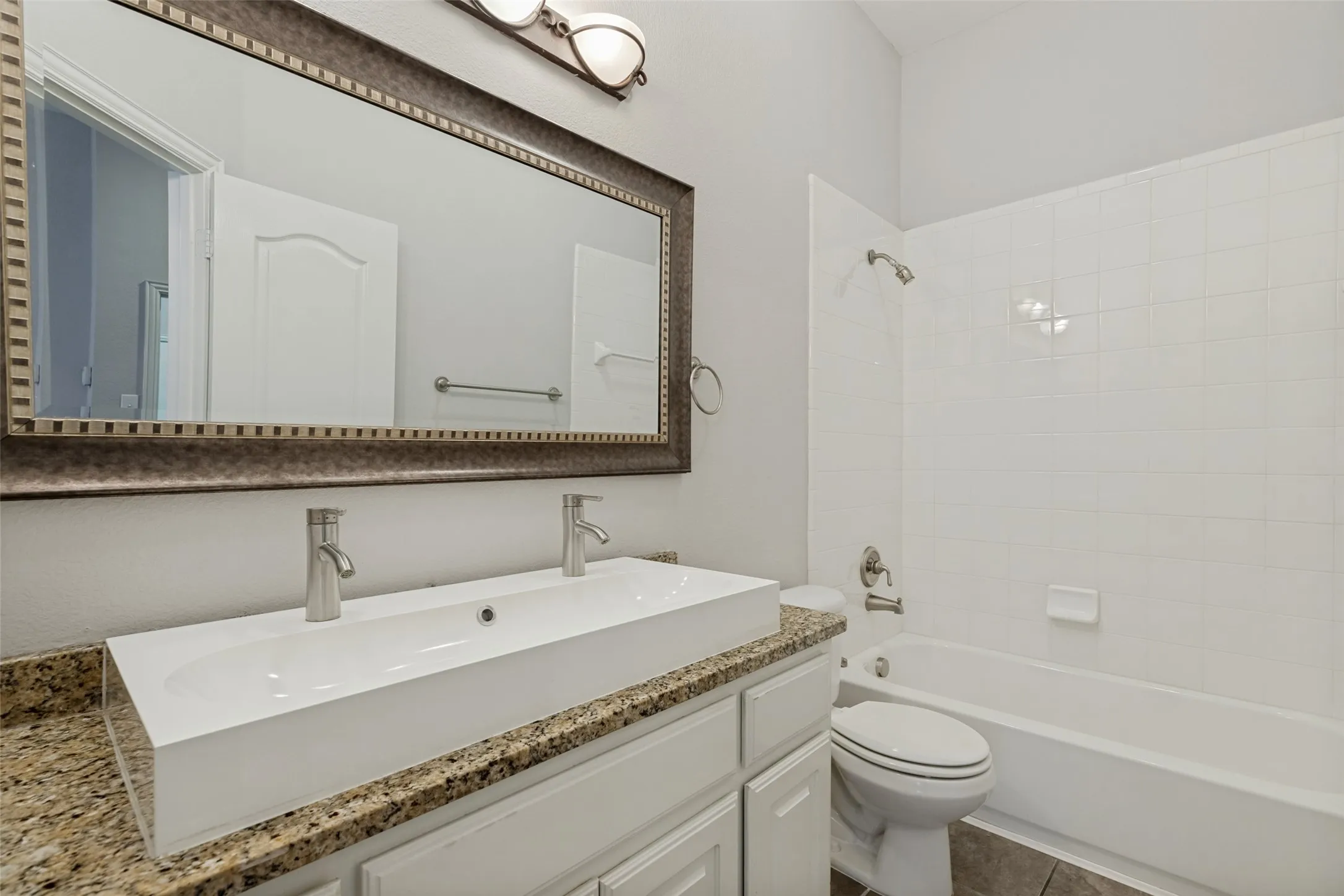 Double vessel sink, granite-style countertop, white cabinetry, large framed mirror, and a full bathtub with tile surround