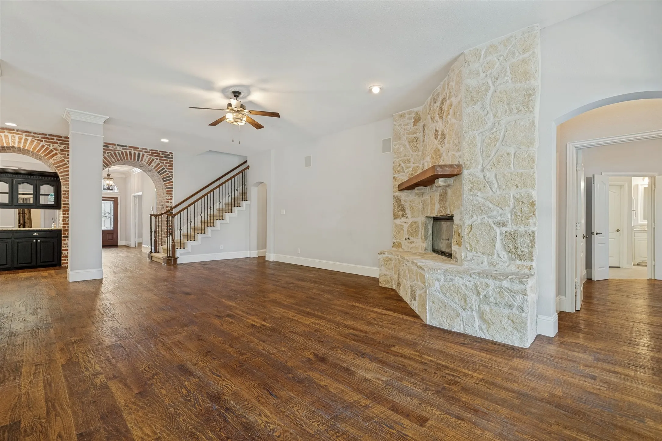 Hardwood flooring, stone fireplace with wood mantel, built-in cabinetry, brick archways, and a staircase with wrought iron balusters