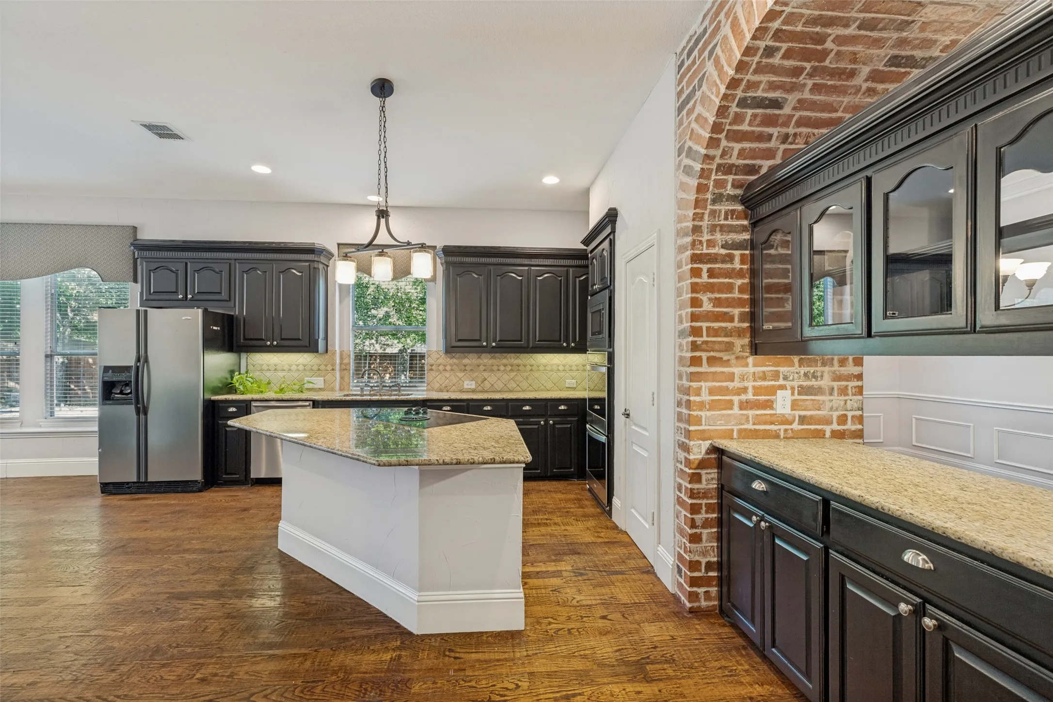 Kitchen featuring dark cabinetry, lighter granite countertops, brick archway, central island, and wood-finish flooring