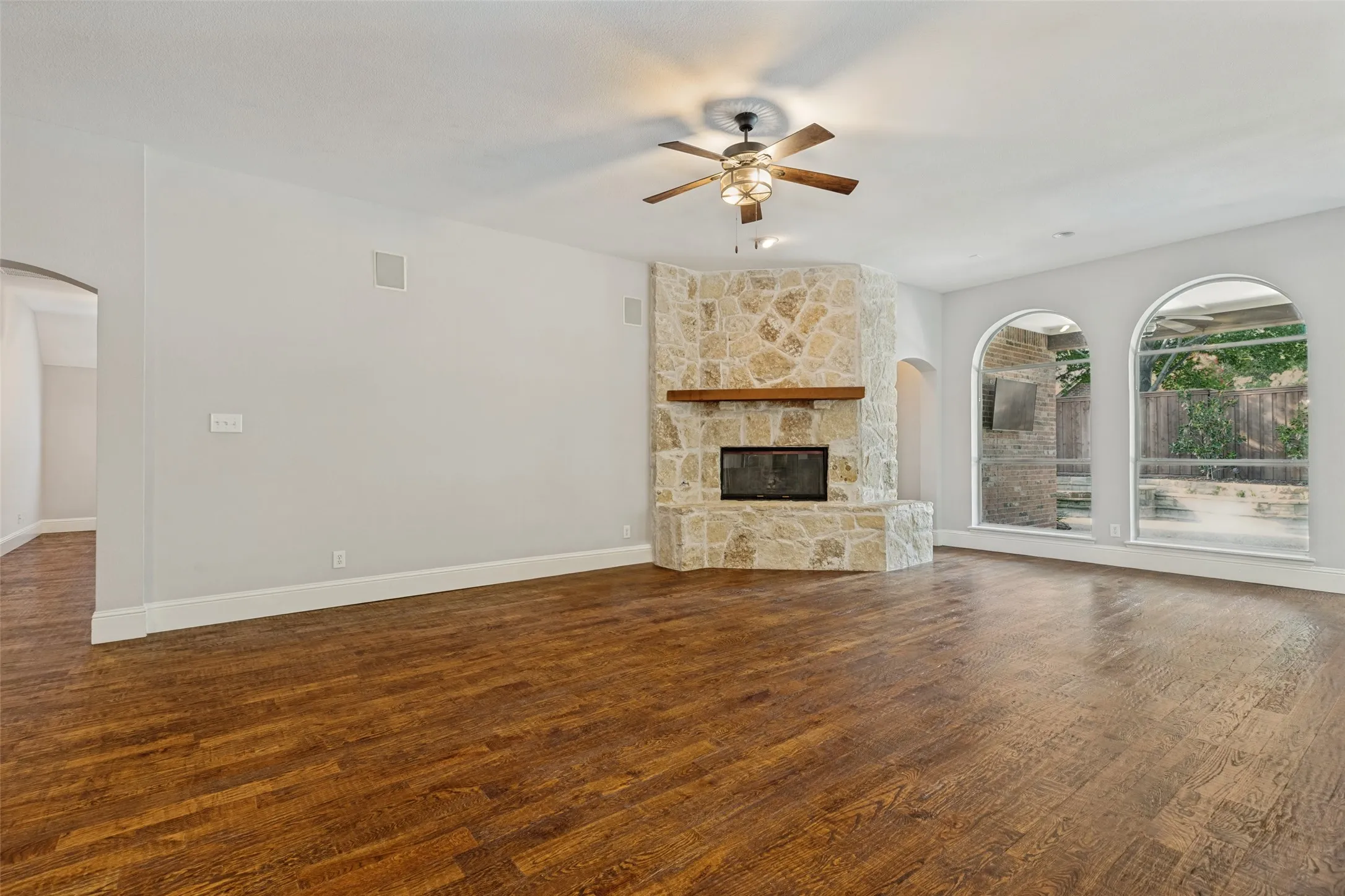 Wood-finish flooring, stone-clad corner fireplace with wood mantel, two arched windows, built-in ceiling speakers, and an arched entryway