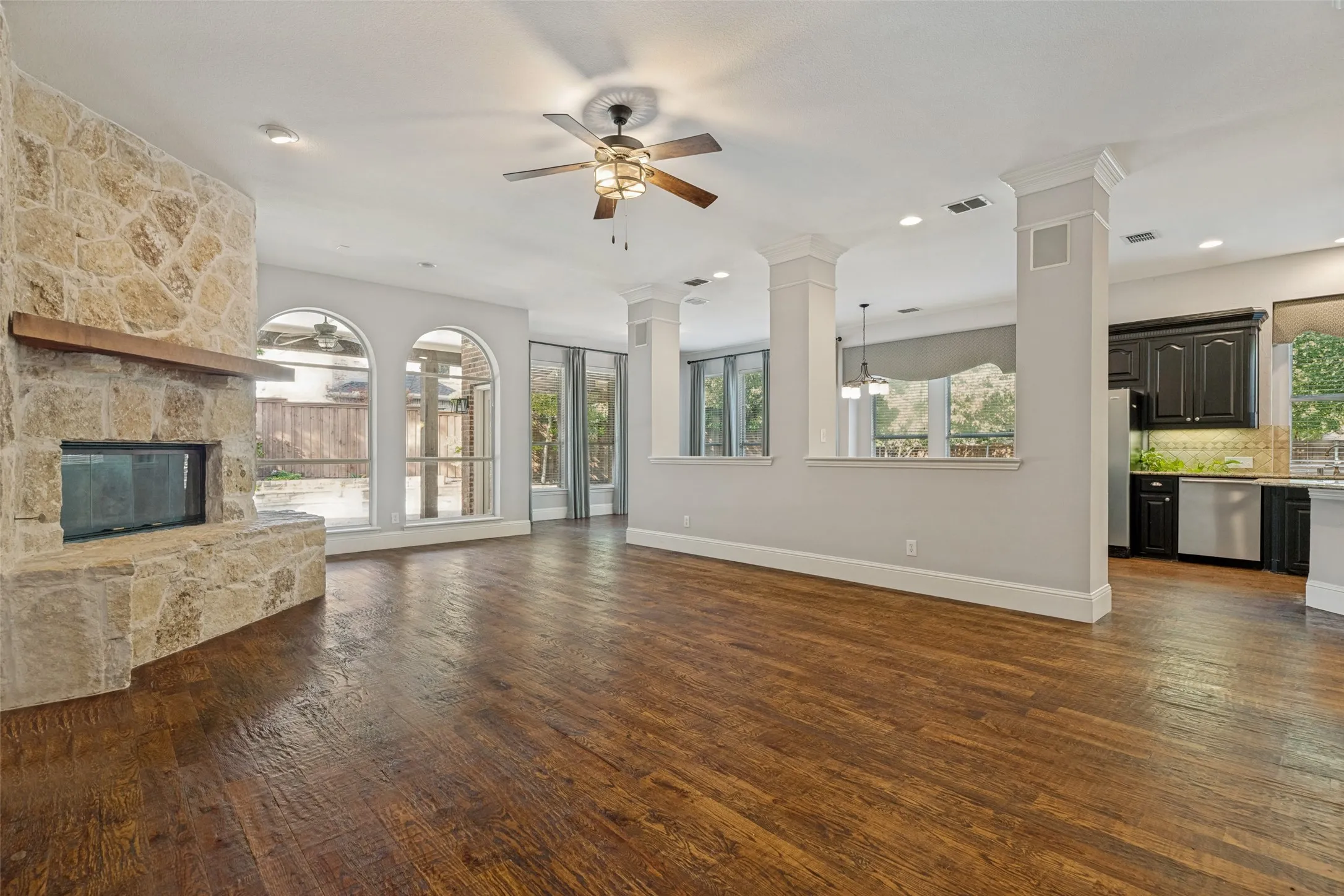 Wood-finish flooring, stone-clad fireplace with wood mantel, arched windows, recessed lighting, and kitchen with dark cabinetry and stainless steel appliances