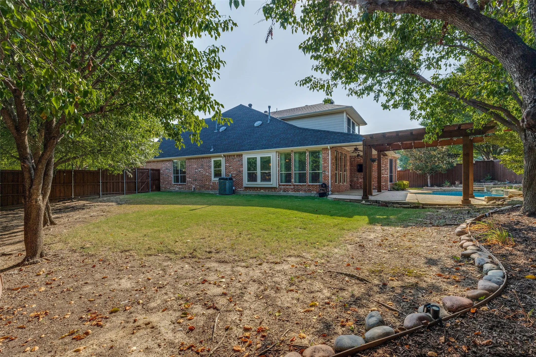 Brick exterior with multiple windows, covered patio with wood pergola, stone-edged landscaping, and a fenced yard with a pool