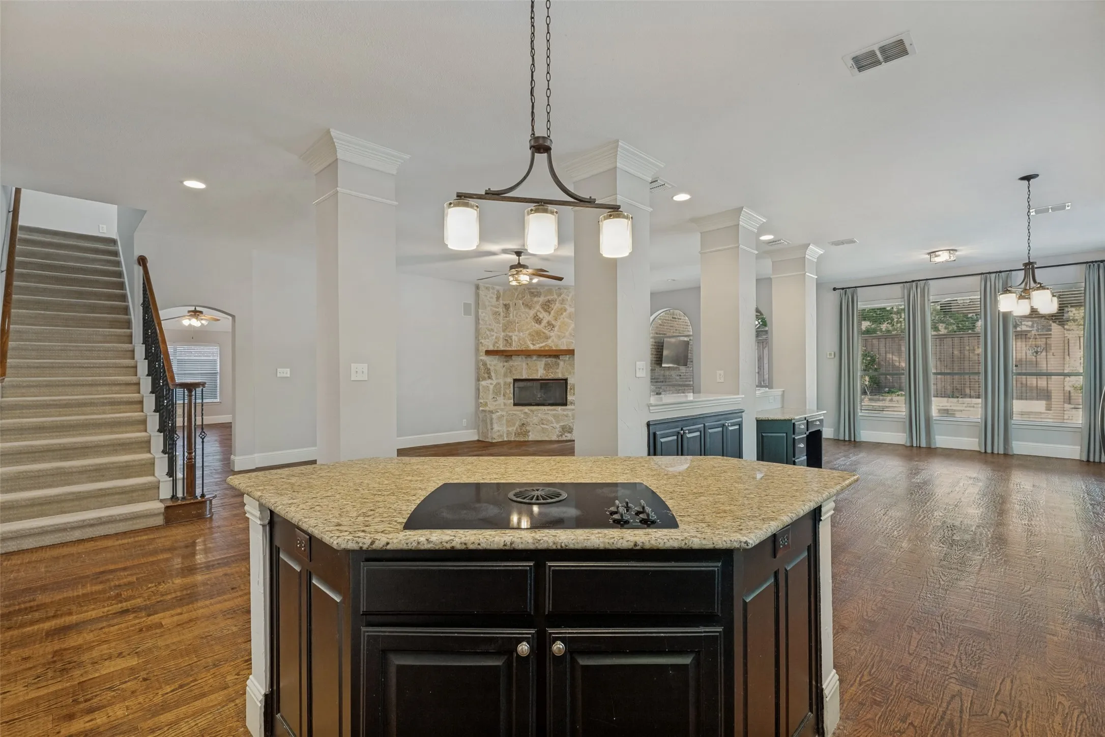 Kitchen island with a built-in cooktop and granite countertop, dark wood-finish flooring, staircase with carpeted treads and wood handrail, stone fireplace surround, and large windows