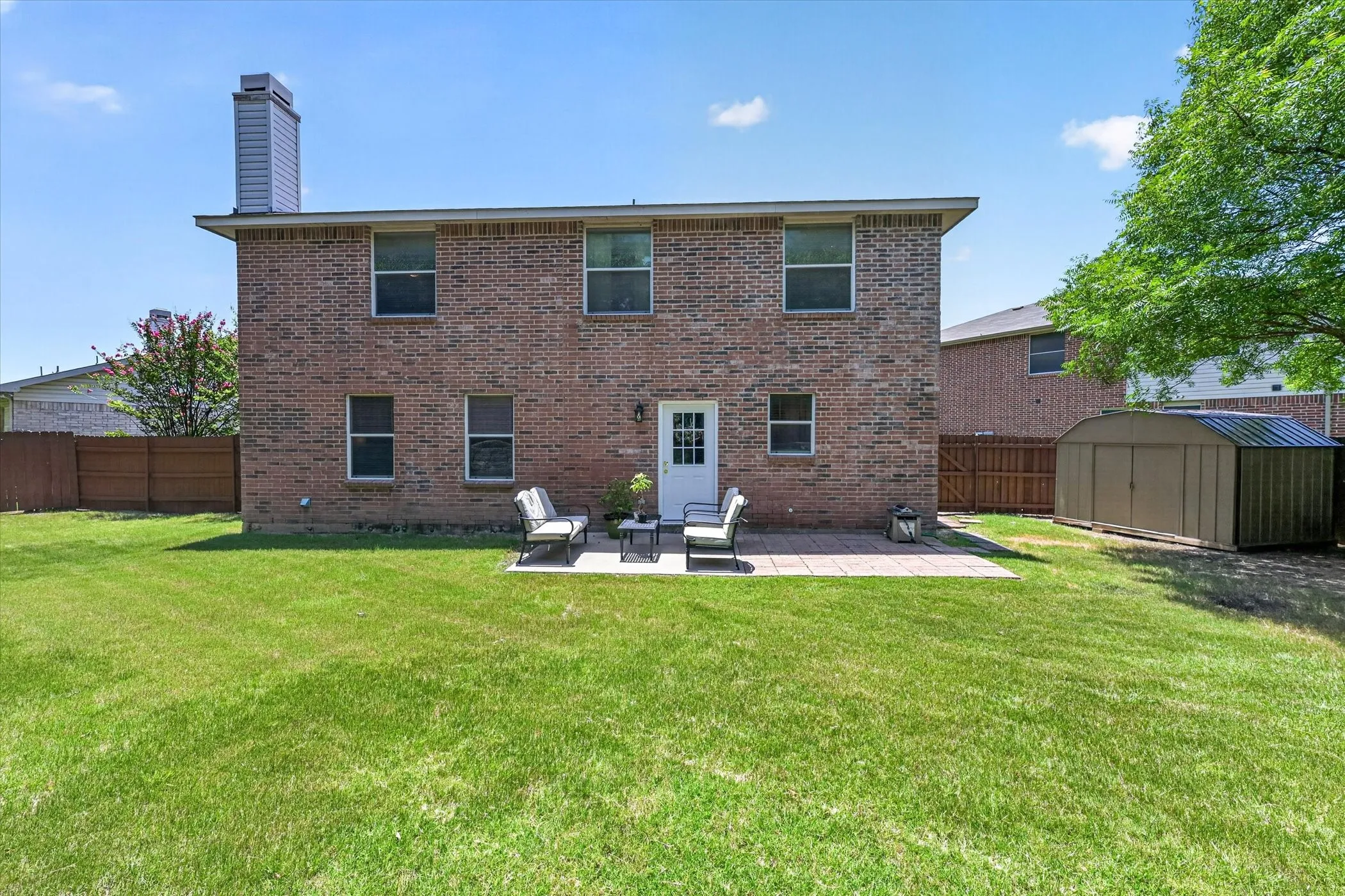Expansive backyard featuring a manicured lawn, brick exterior, rectangular patio, white rear door with glass panels, and a metal storage shed