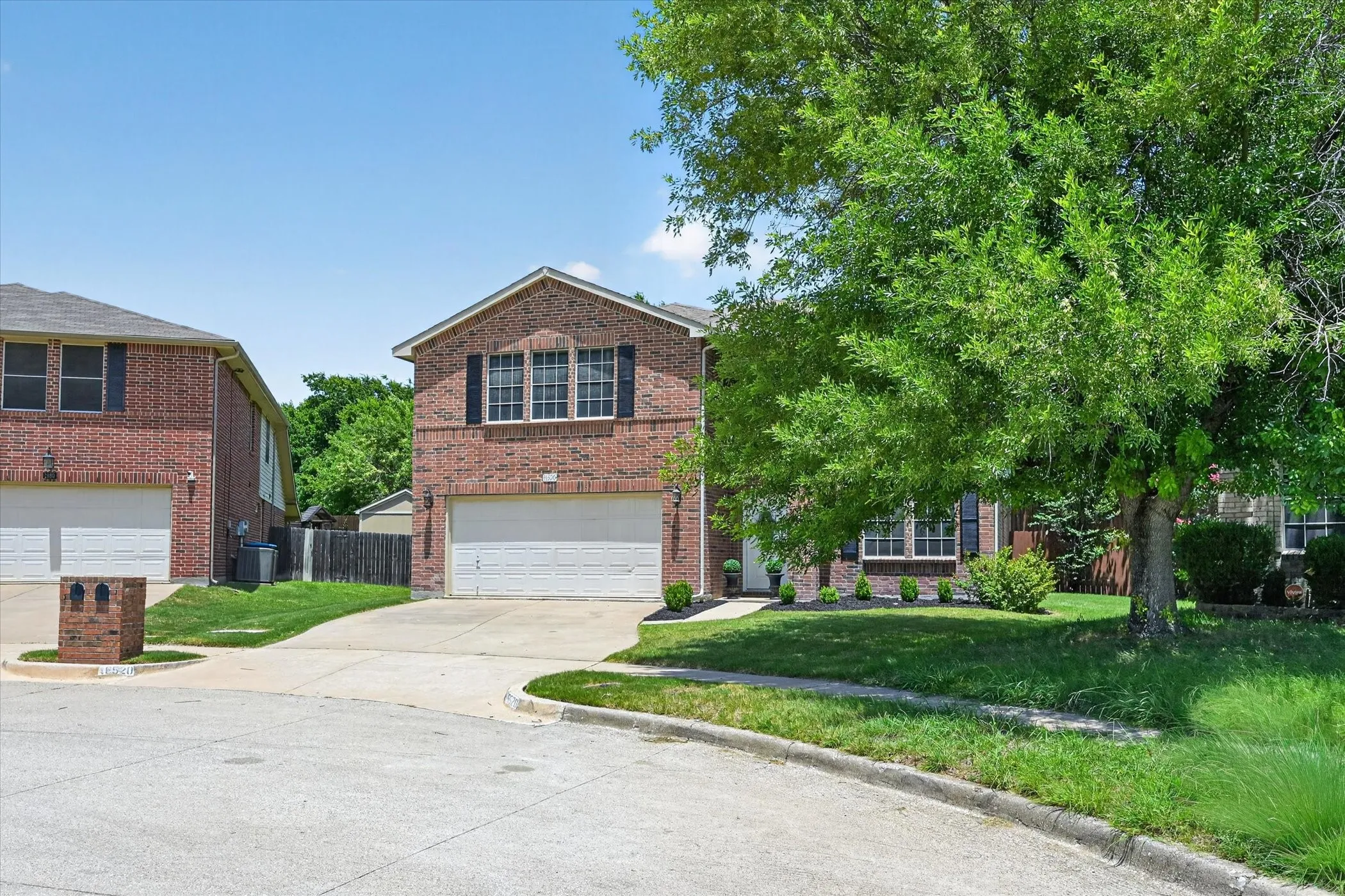 Brick facade two-story residence featuring a two-car garage, mature front yard tree, and manicured landscaping