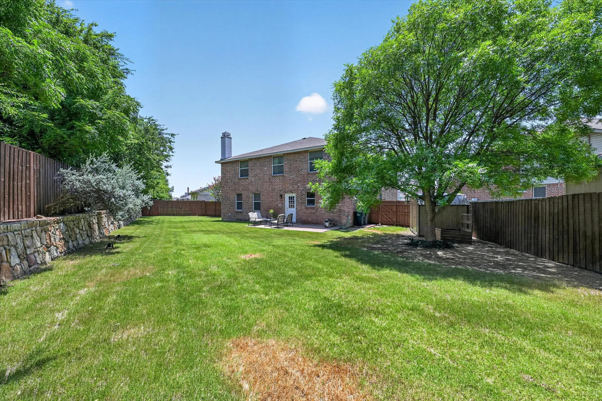 Spacious backyard featuring a large brick two-story residence, a stone retaining wall, a mature shade tree, privacy fencing, and a concrete patio