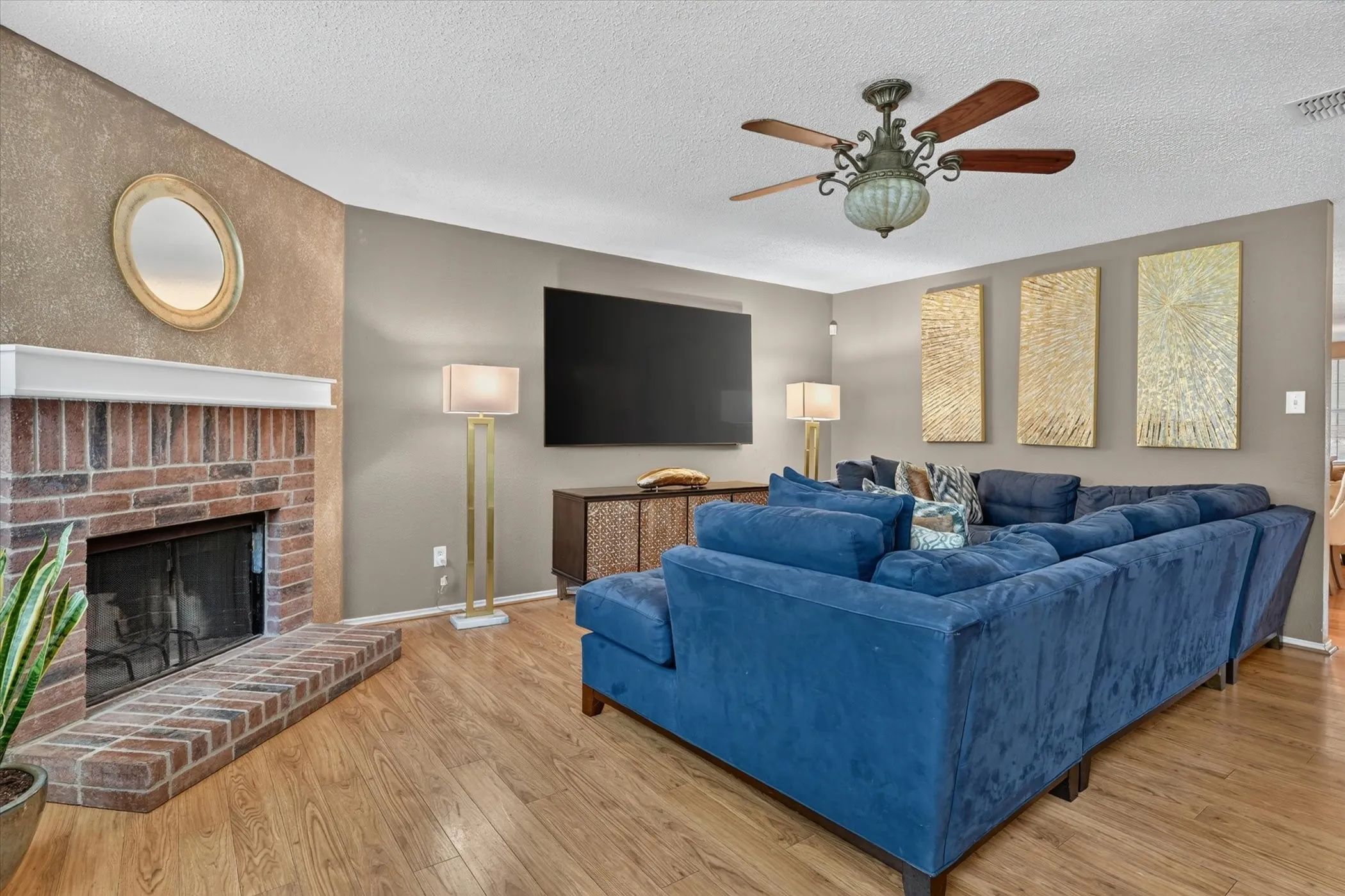 Brick fireplace with a white mantel, wood-finish flooring, textured wall accent, ceiling fan, and built-in entertainment console
