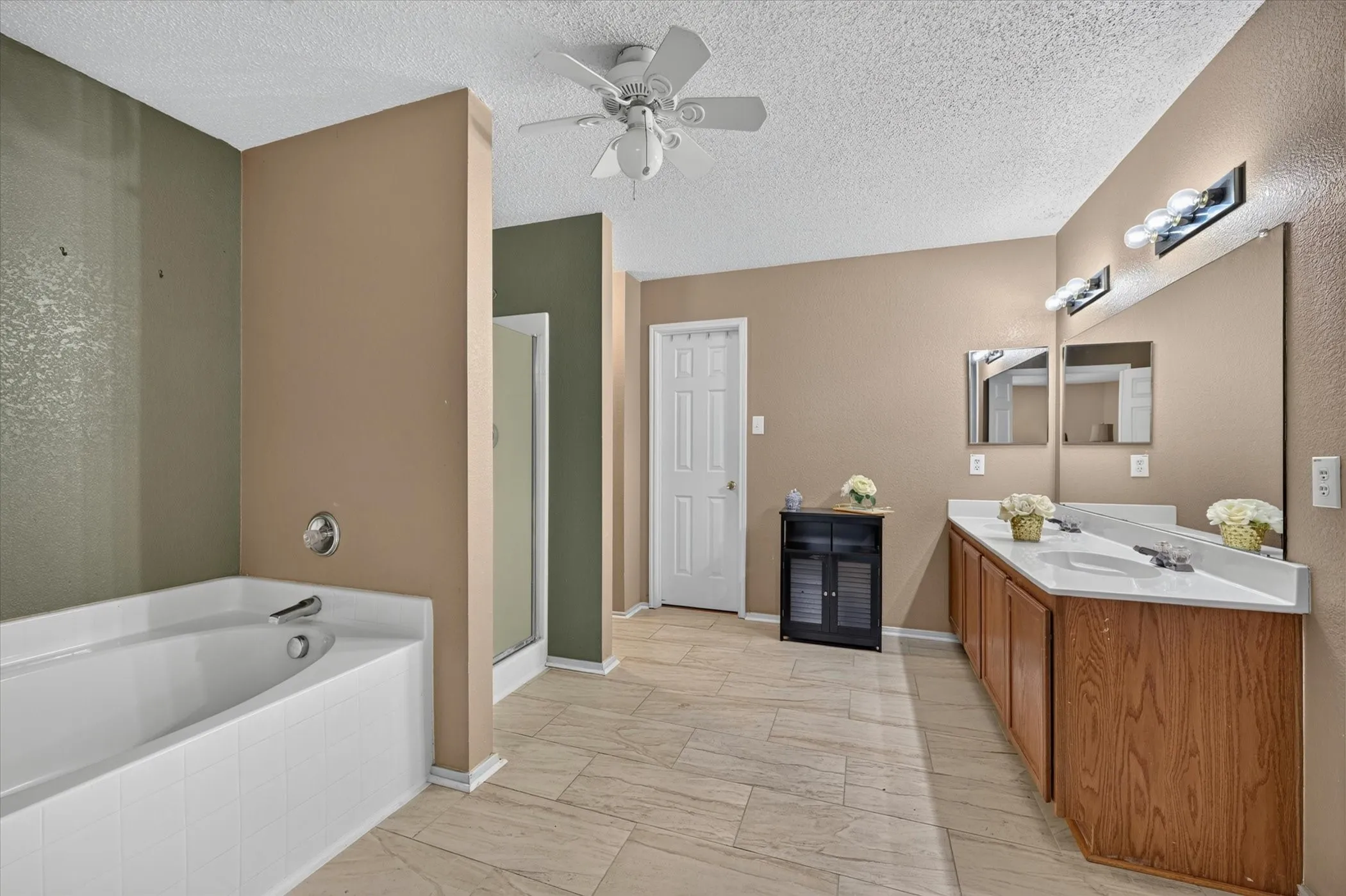 Bathroom featuring a built-in tub, separate shower enclosure, dual vanity with wood-finish cabinetry, neutral wall paint, and ceiling fan