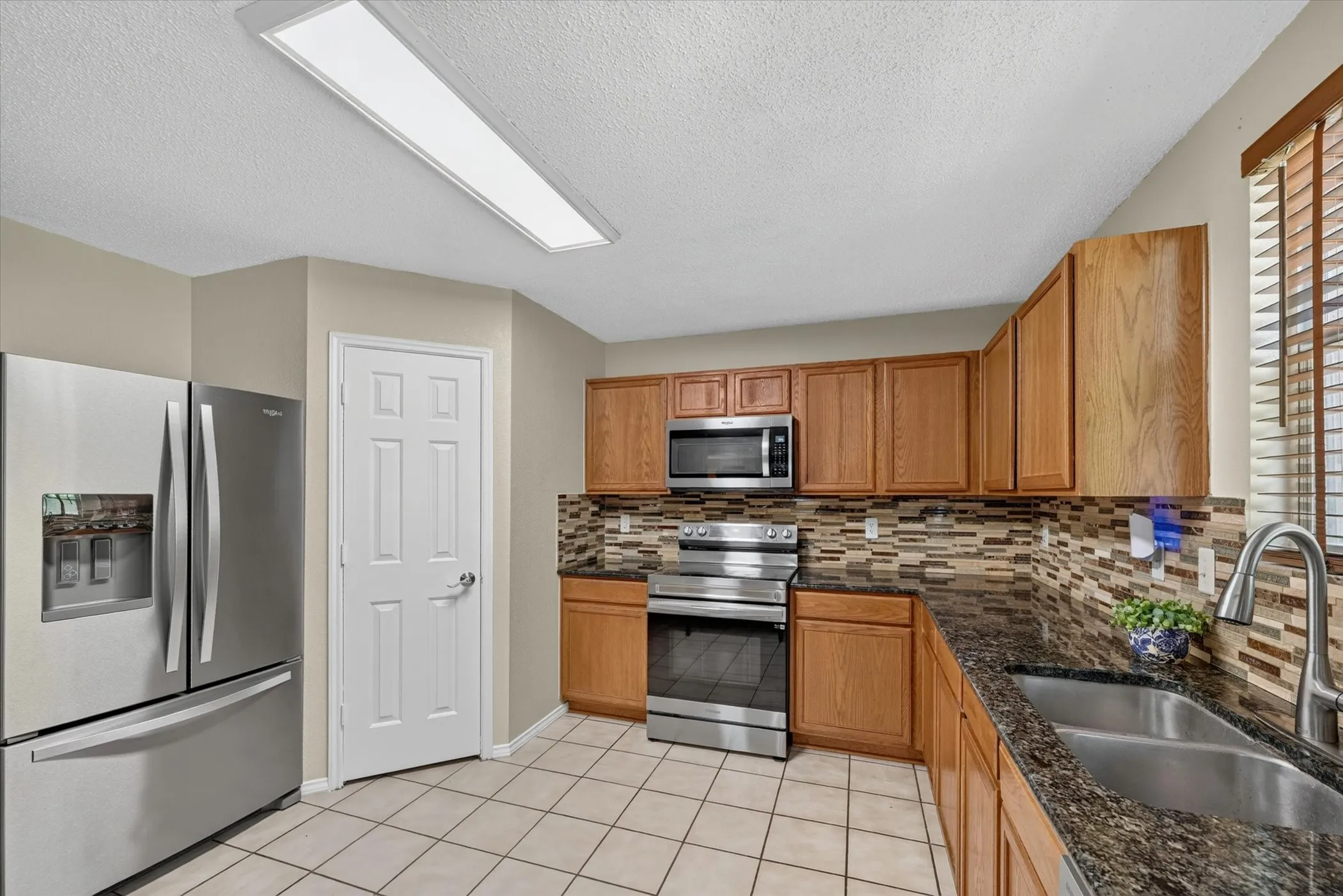 Kitchen featuring stainless steel appliances, wood-finish cabinetry, granite-style countertops, mosaic tile backsplash, and ceramic tile flooring